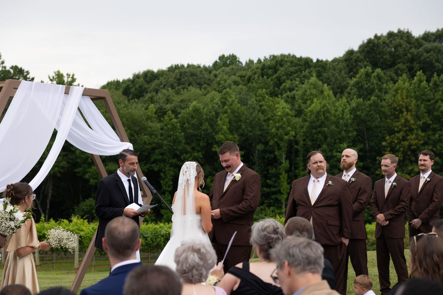 An outdoor wedding ceremony takes place under white fabric draped between trees with guests seated on white chairs.