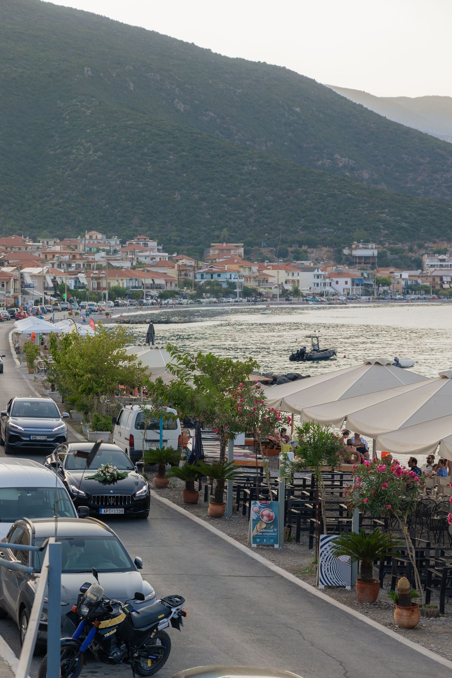 Coastal town view with mountains, parked vehicles, and a scenic harbor with boats in the distance.