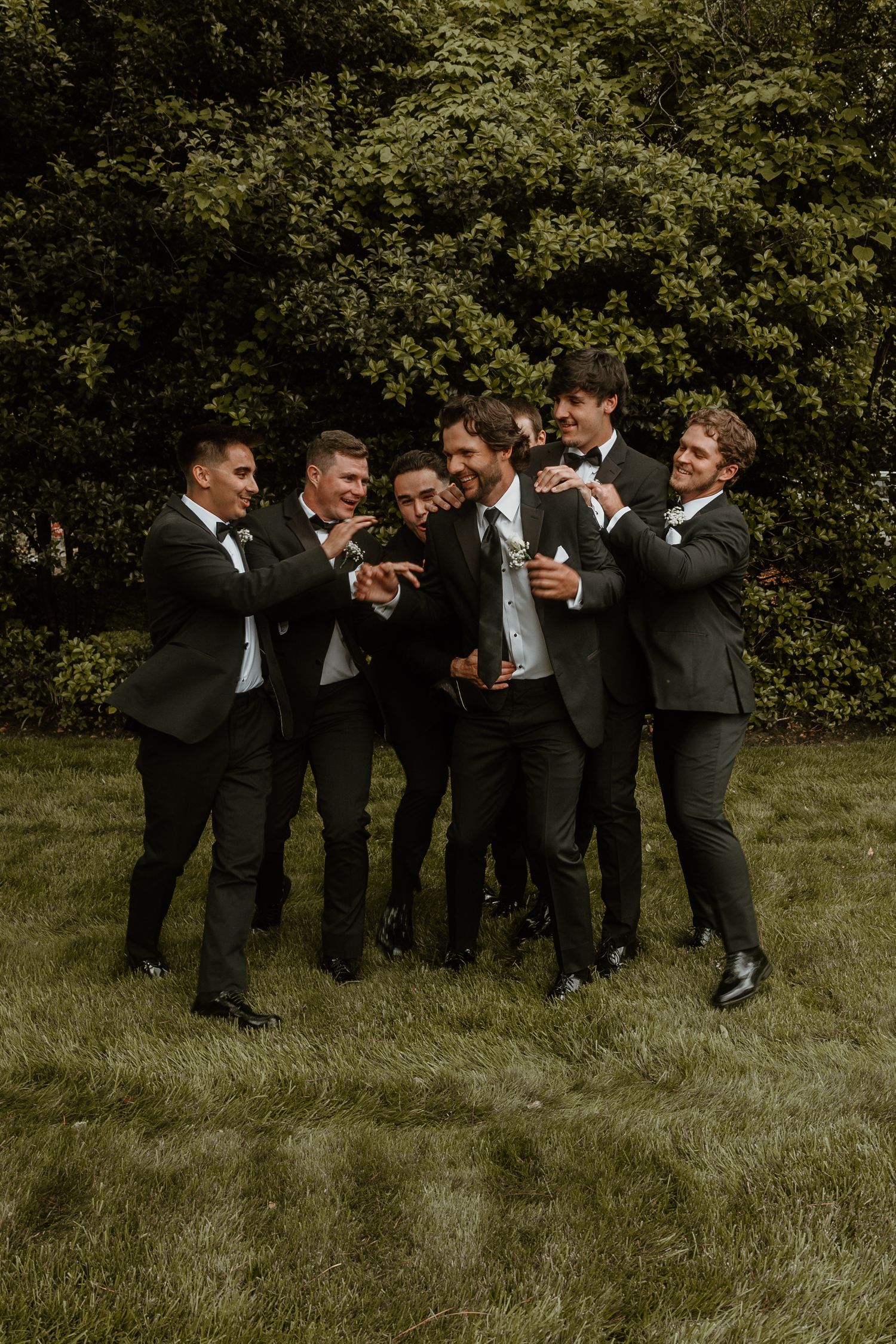 Groomsmen in black tuxedos laughing together on a grassy field.