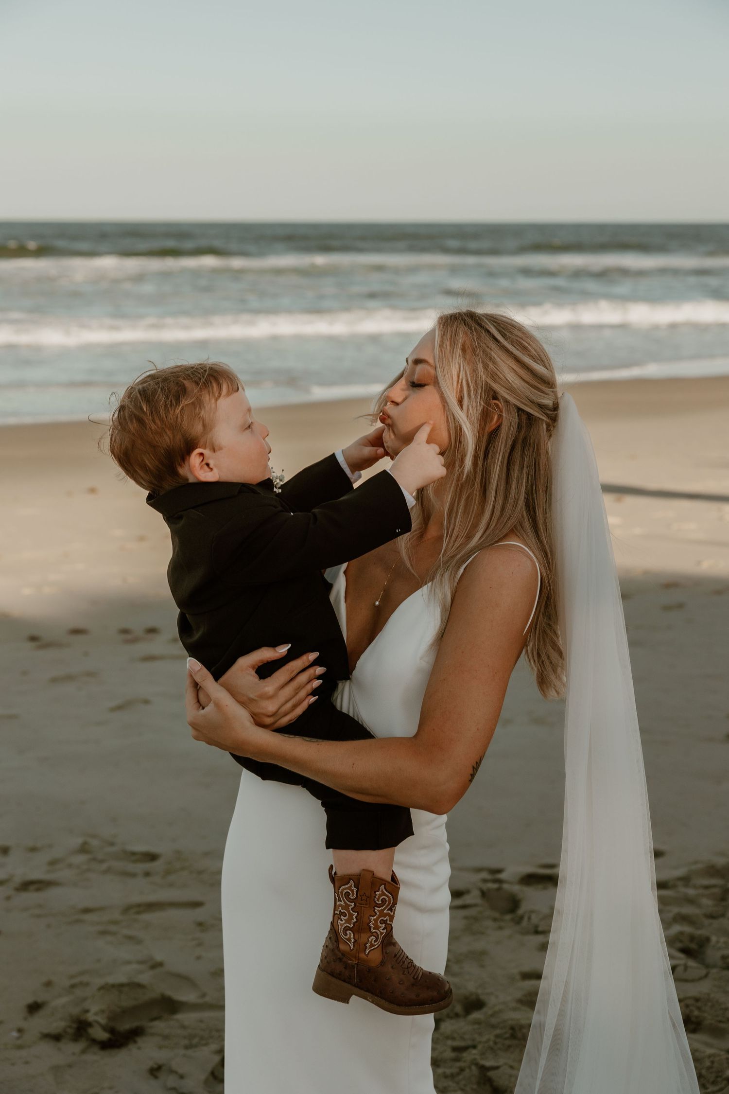 Sweet moment between bride in white gown and small child on beach at sunset.