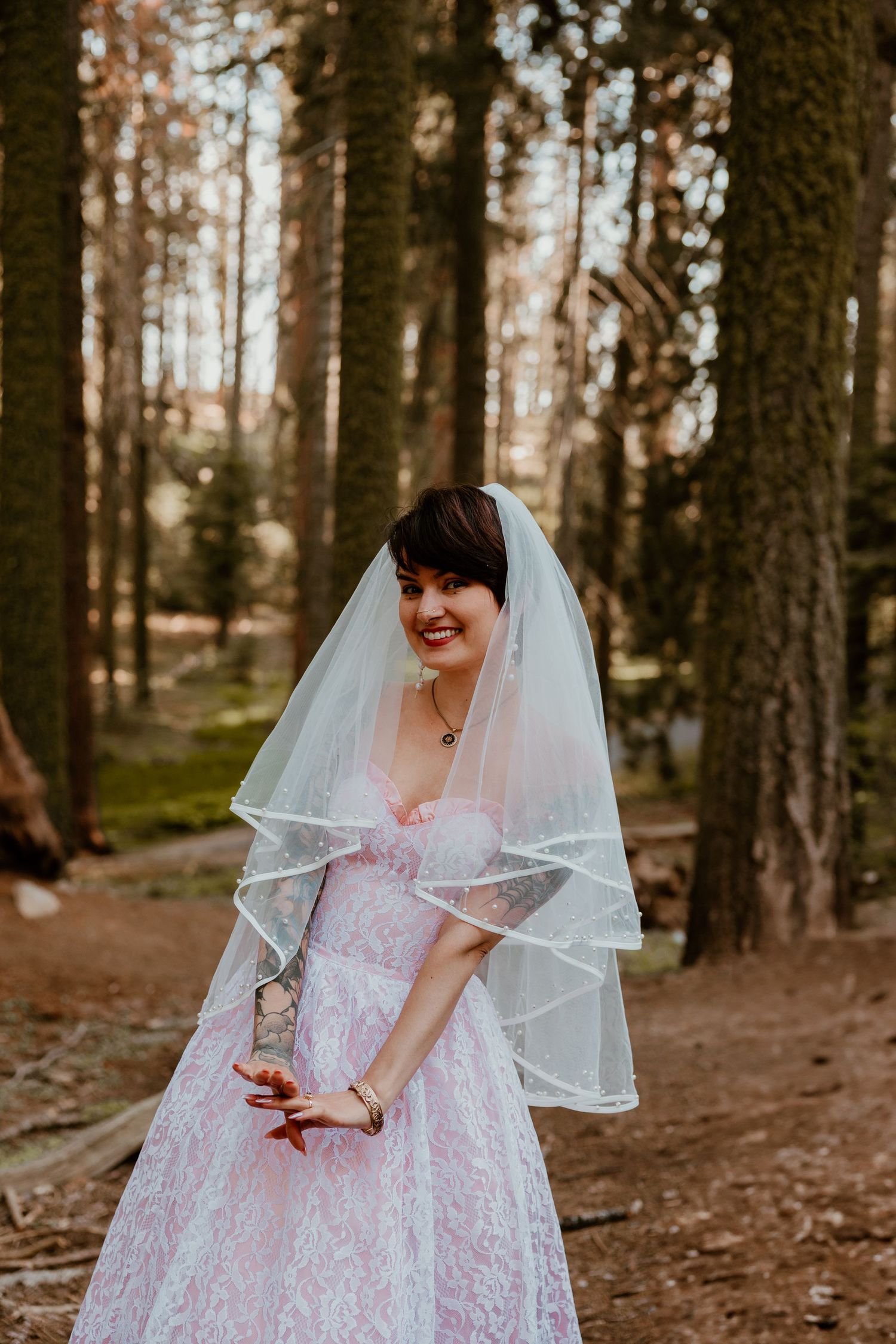A vintage-inspired wedding scene in a forest with a pink lace dress and flowing veil.