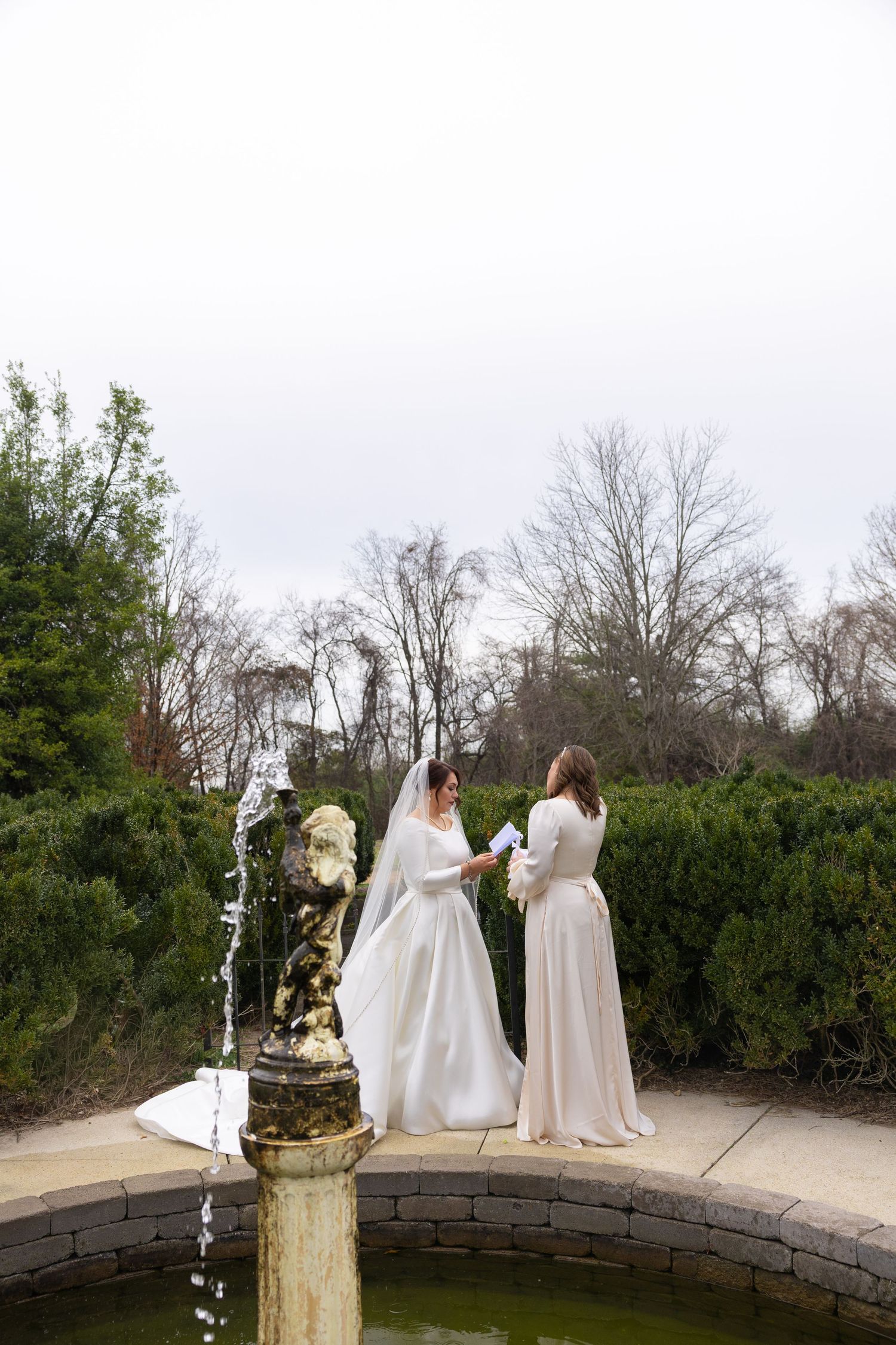 Two figures in flowing white dresses stand together on a curved stone wall overlooking a garden landscape.
