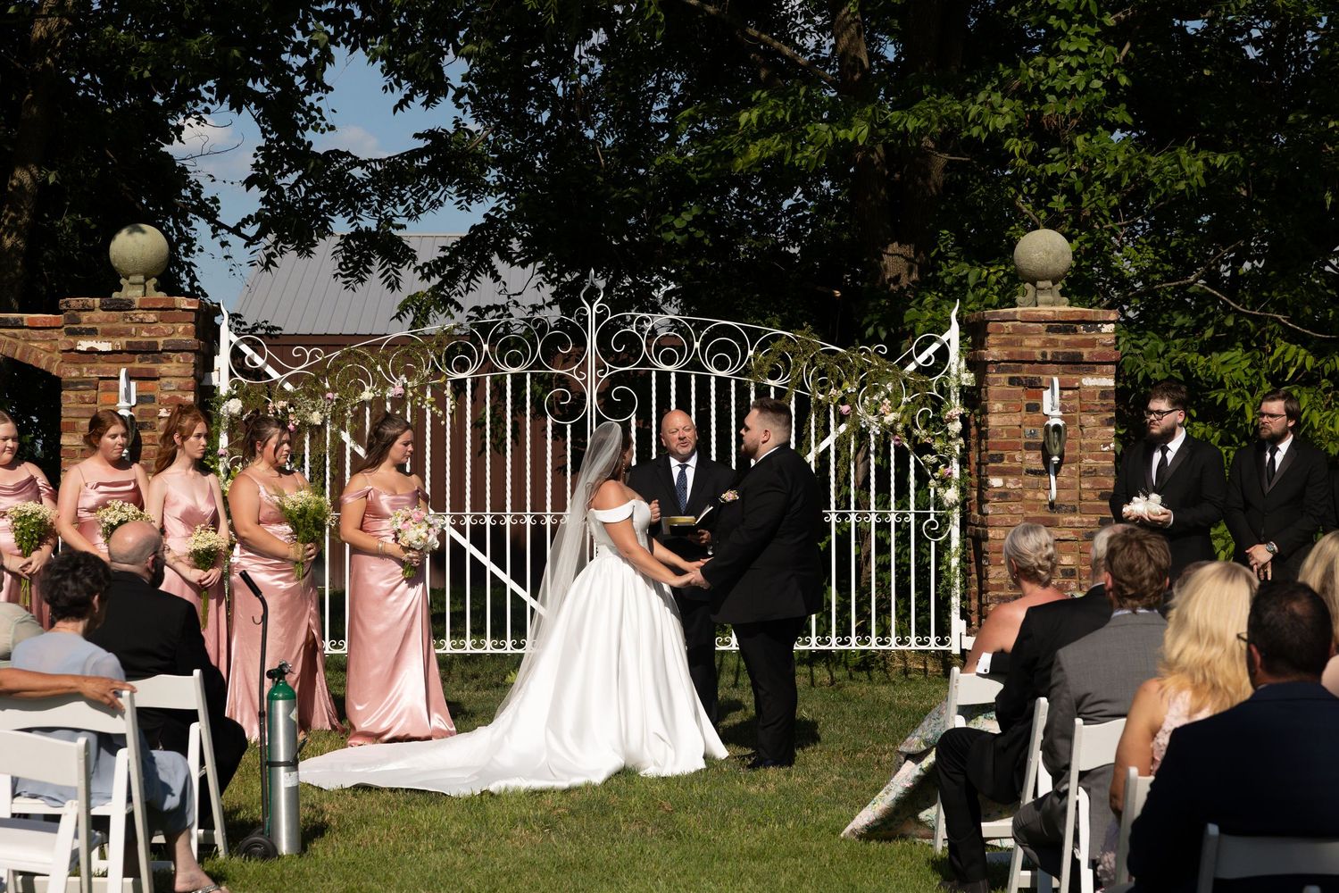 Wedding party members walk along a stone wall and iron gate during an outdoor celebration.