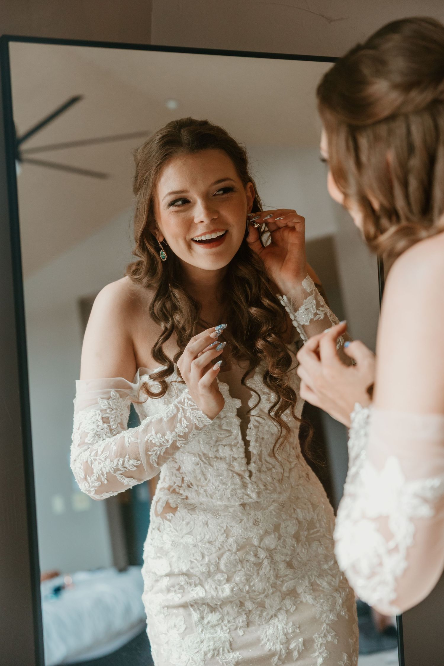 A person in a wedding dress puts on earrings while looking in a mirror.