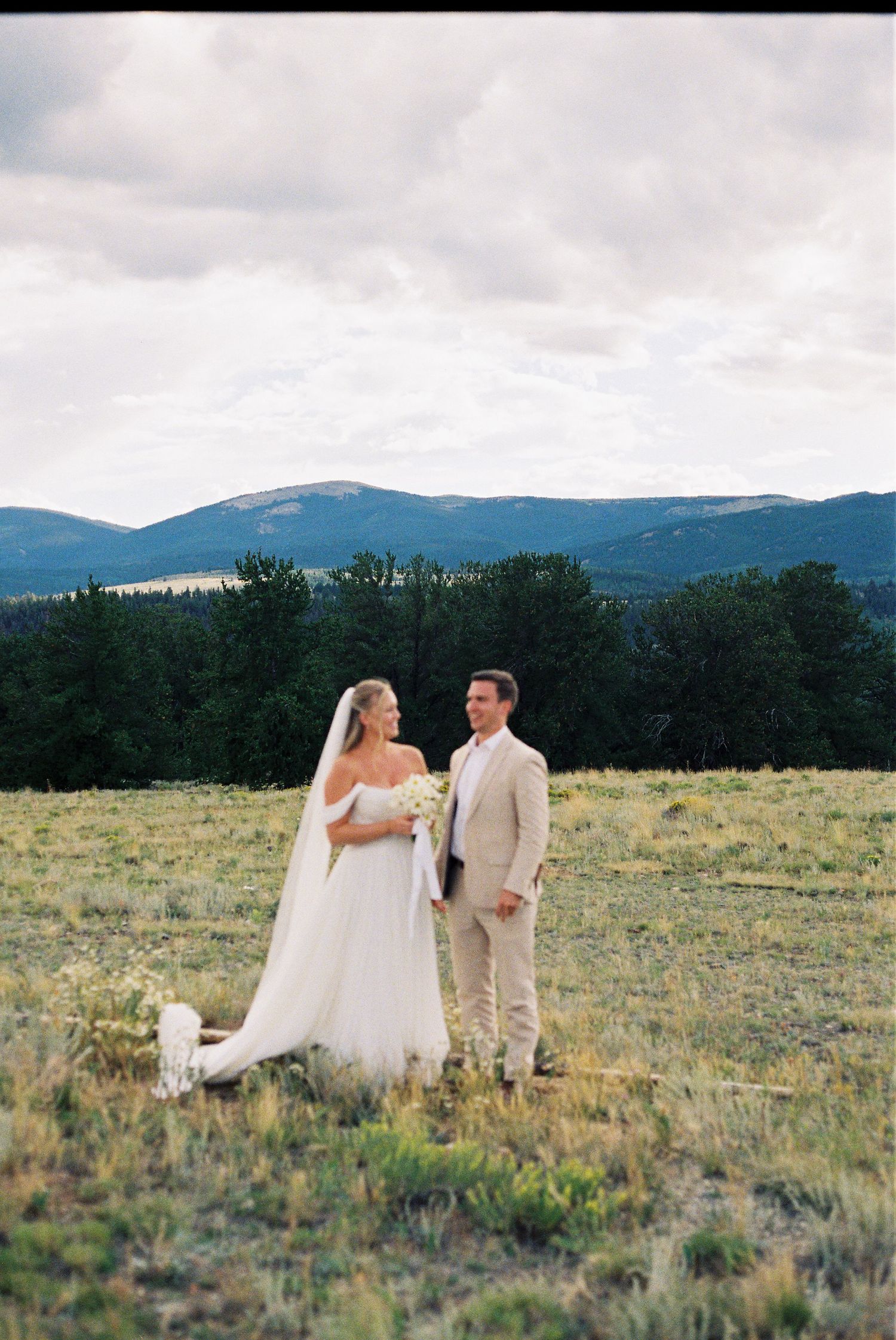 Couple poses in a scenic mountain meadow with evergreen forest and peaks in the background.