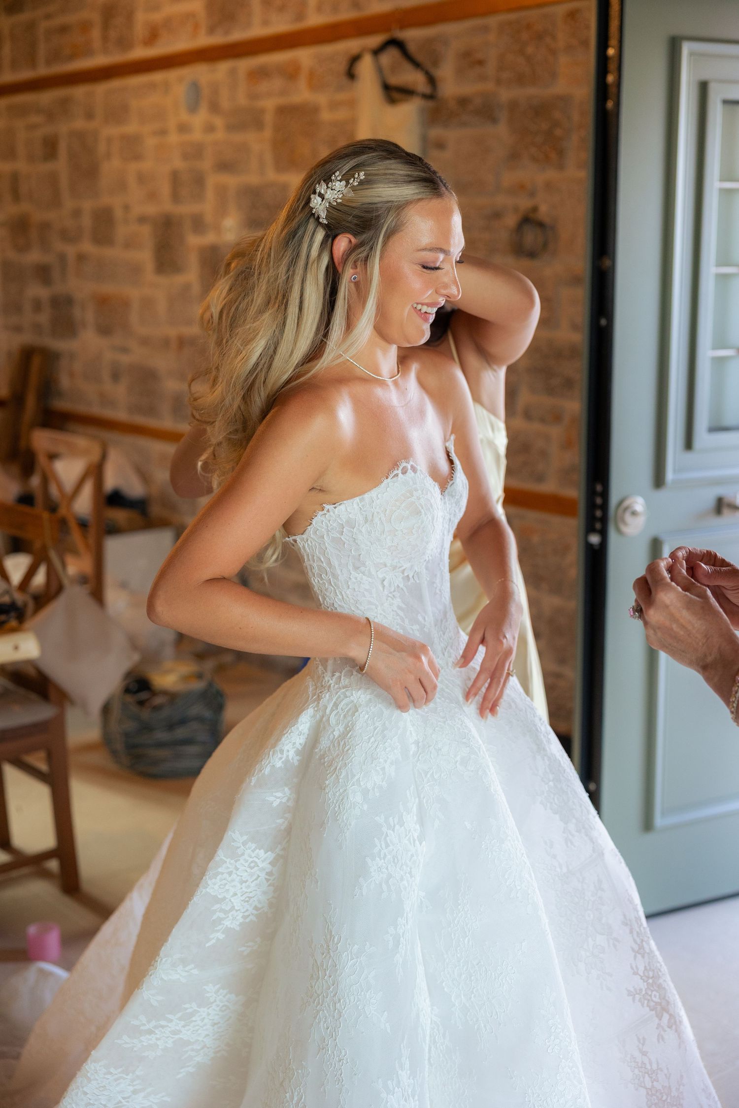 A bride in a white lace ball gown twirls and laughs joyfully while getting ready in a rustic brick-walled room.