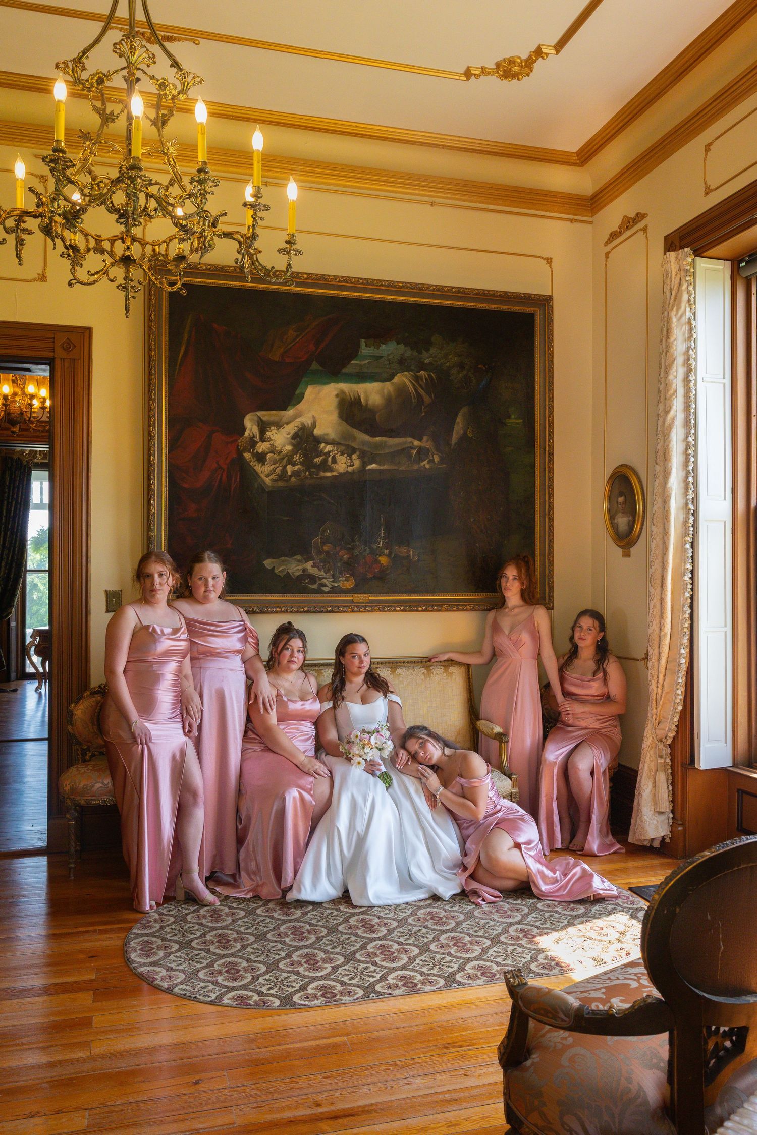 An elegant wedding party poses in a historic room with hardwood floors and a grand chandelier.