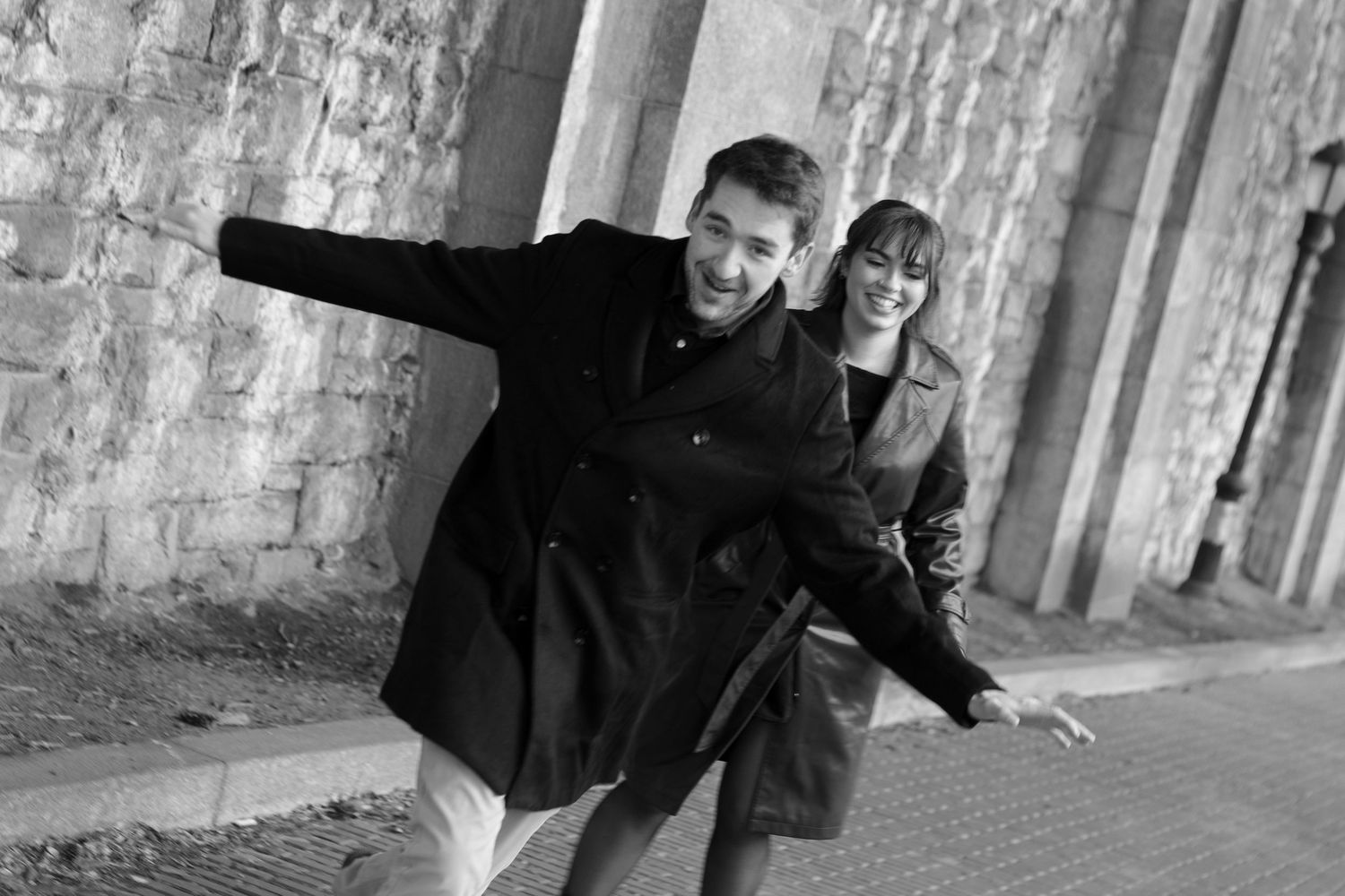 Black and white photo of two people playfully posing under stone archways in an urban setting.