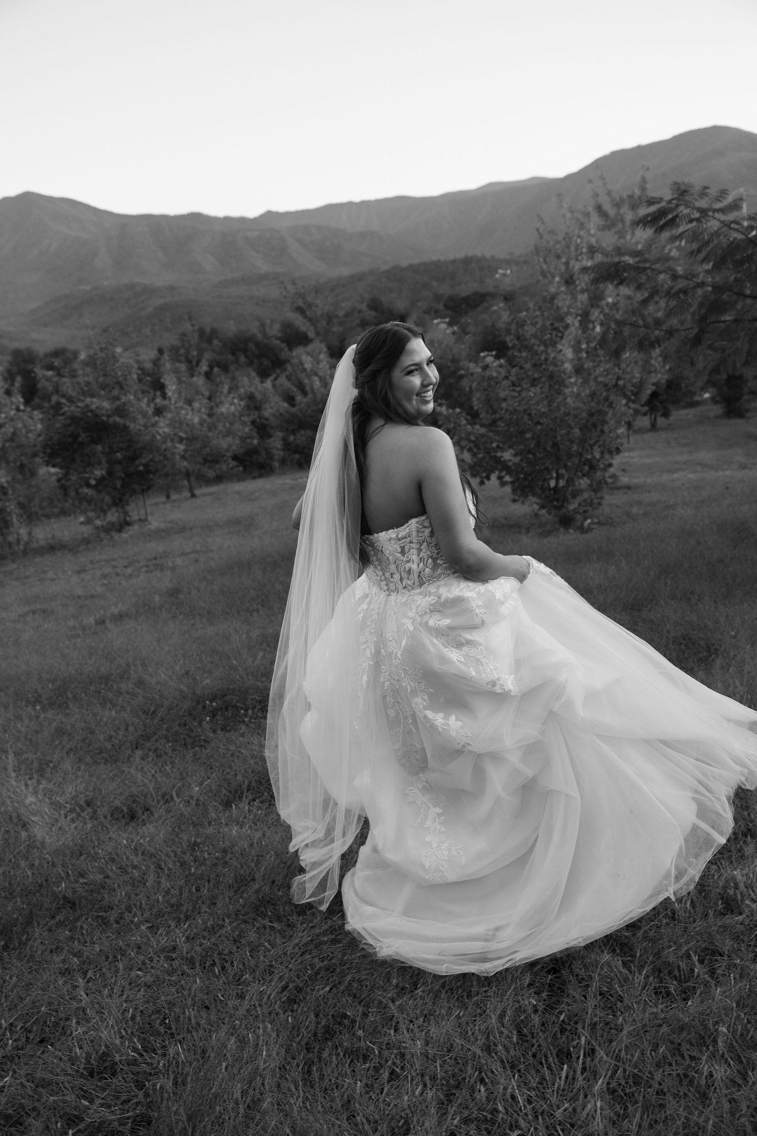Black and white photo of bride in flowing dress twirling against mountain landscape.