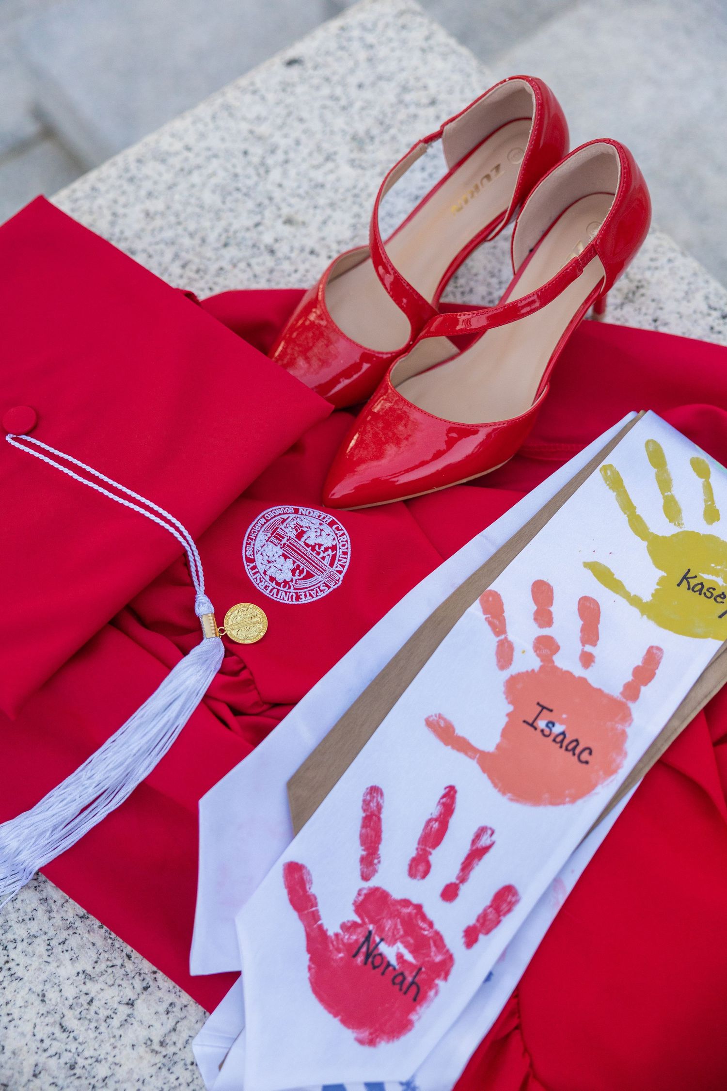 Red graduation gown, cap, red heels, and handprint stole with names displayed on stone steps.