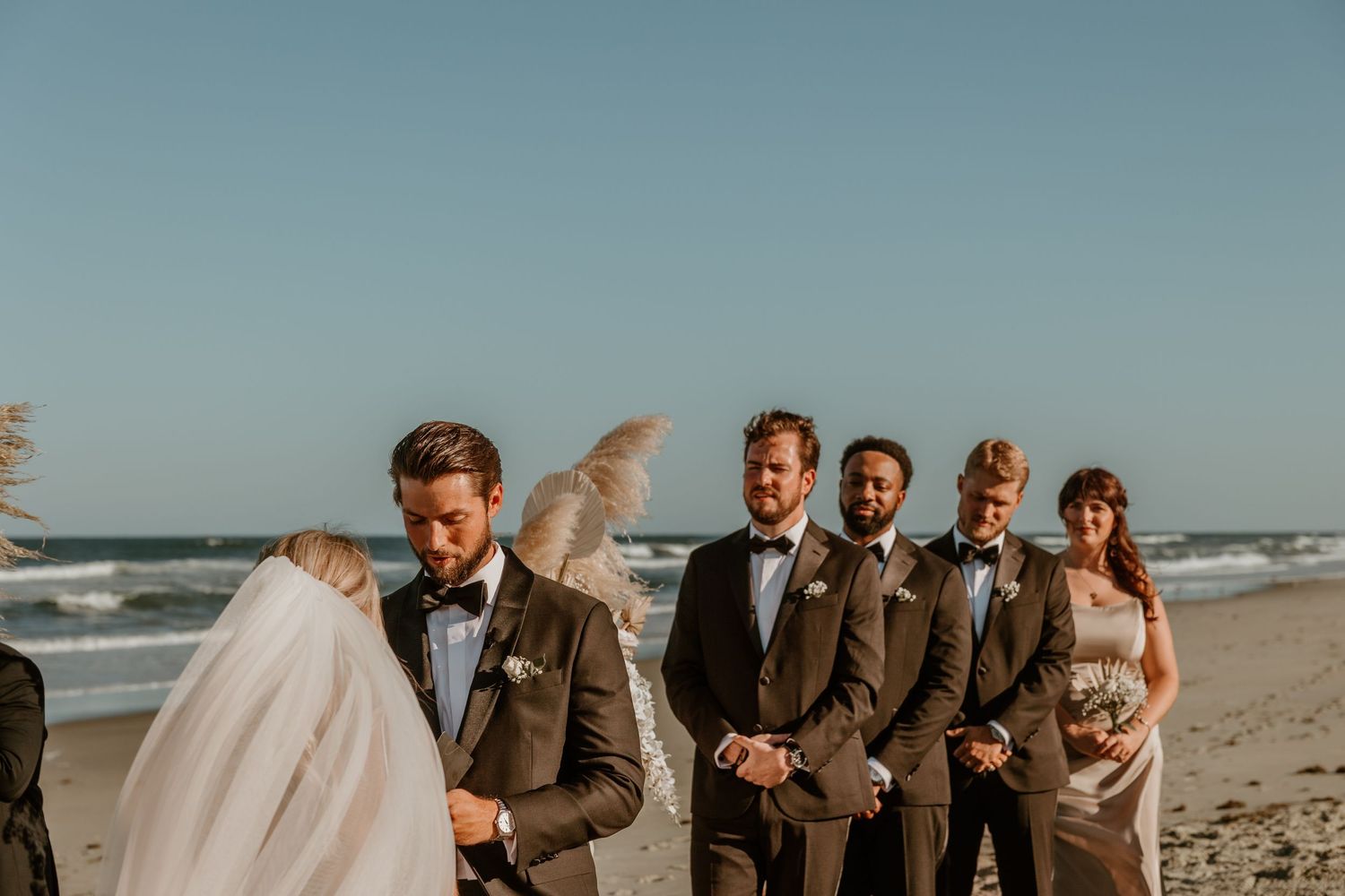 Wedding party gathered on beach with ocean waves and blue sky in background.