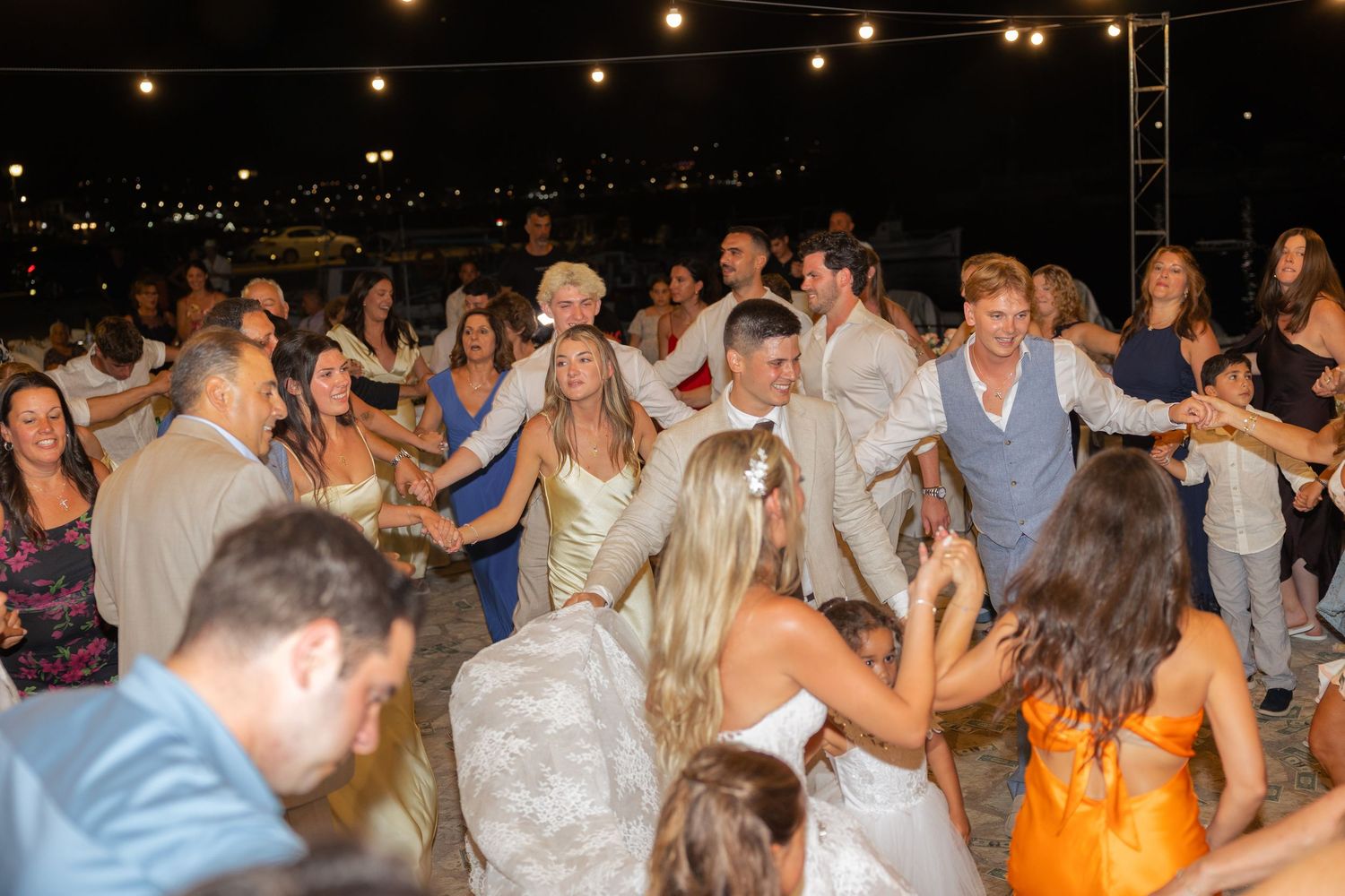 A group of people dancing together at a lively nighttime outdoor party with string lights overhead.