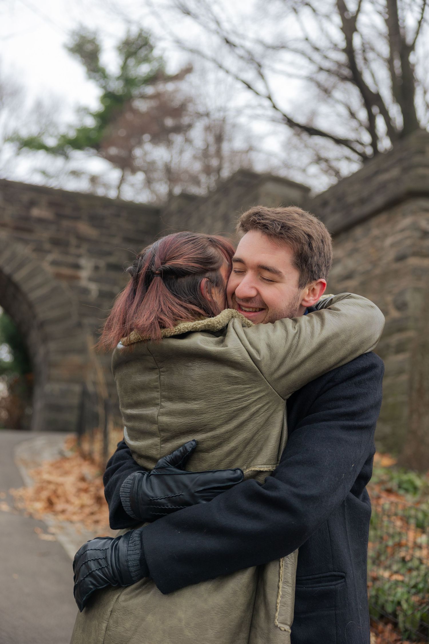 Two people share a tender embrace outdoors near a stone wall on an autumn day.
