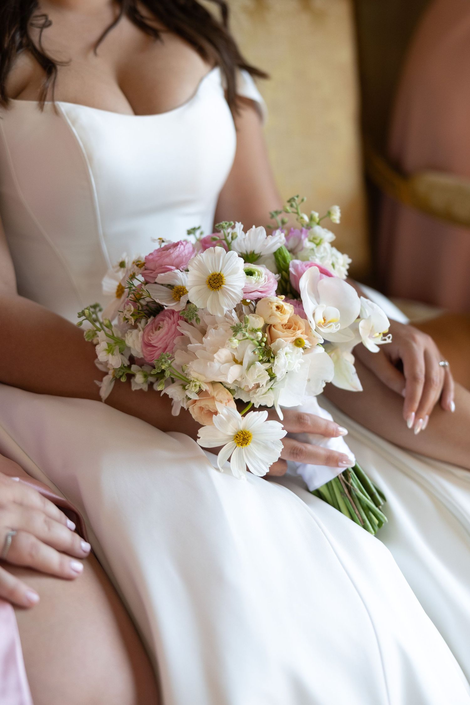 A delicate bridal bouquet with white daisies and pink flowers rests on a white wedding gown.