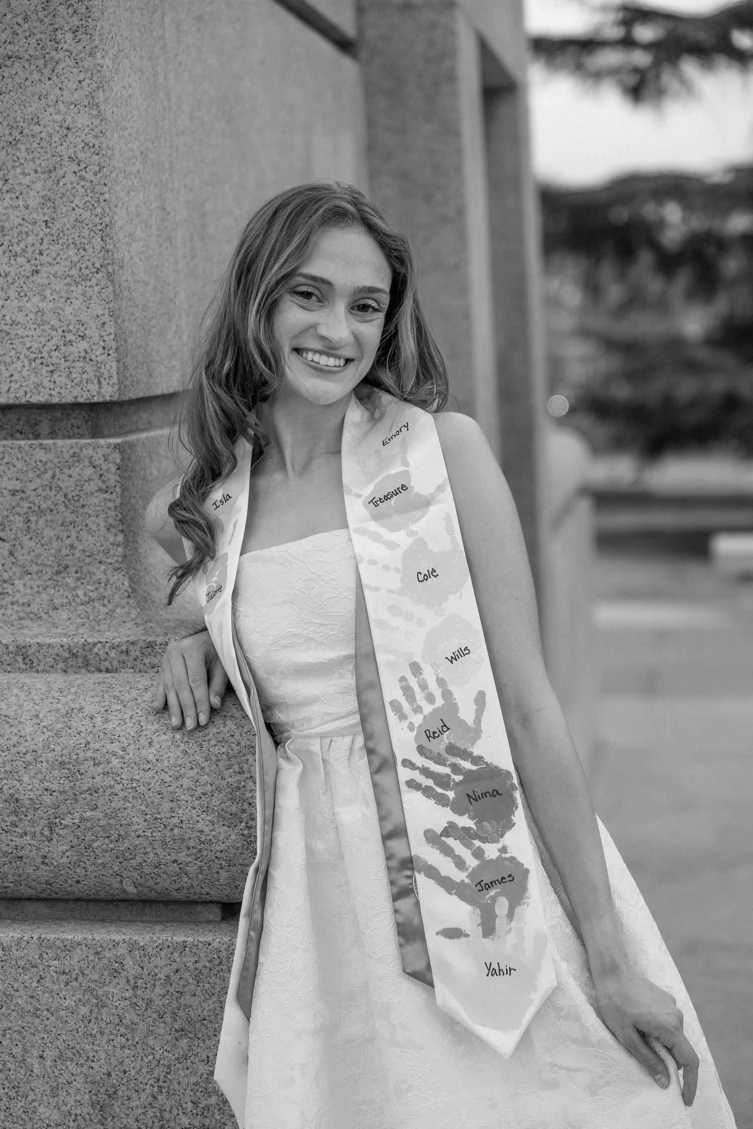 Smiling young woman in white dress wearing a graduation sash with handprints, leaning against a stone column.