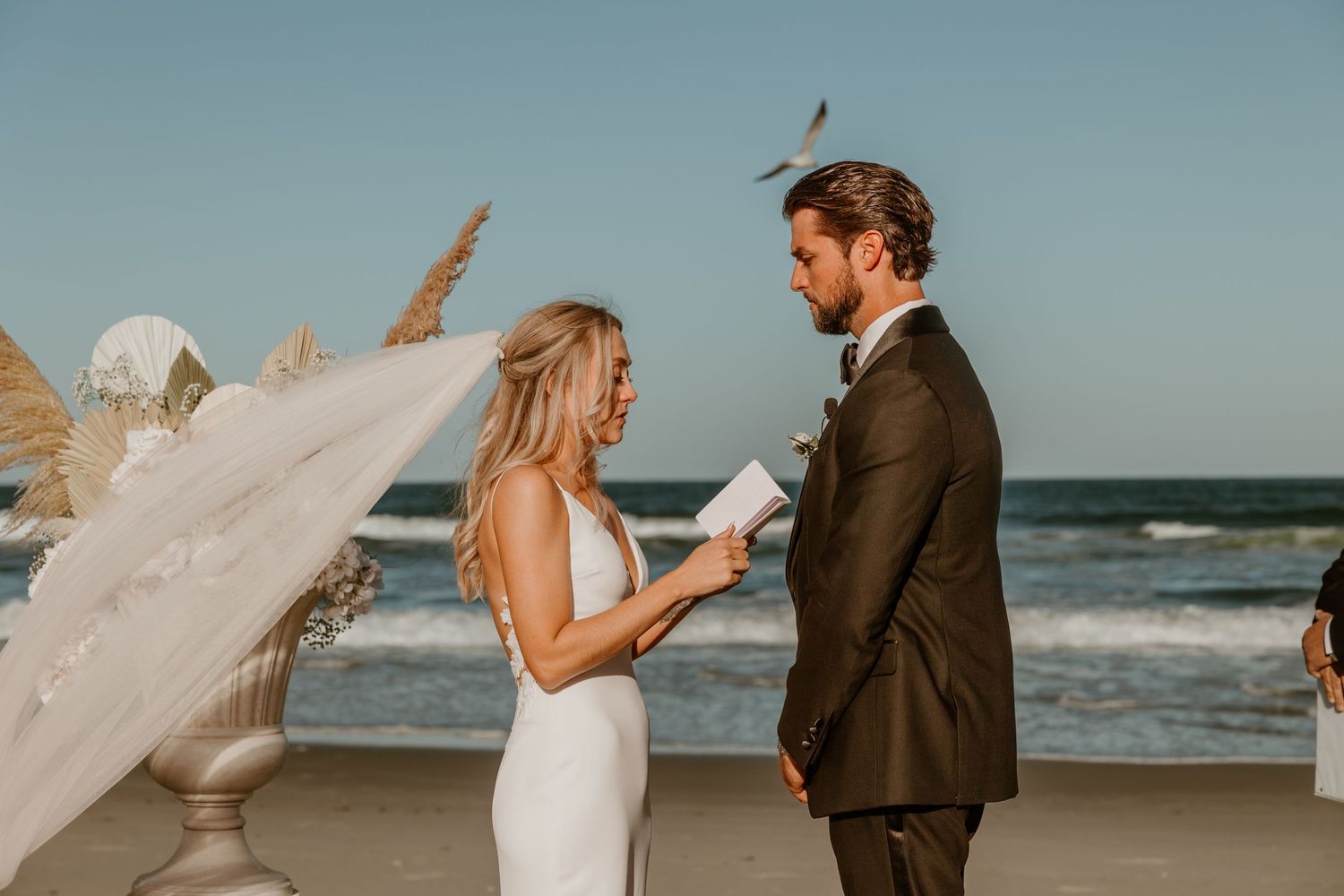 Bride and groom exchanging vows on sunny beach with flowing veil in ocean breeze.