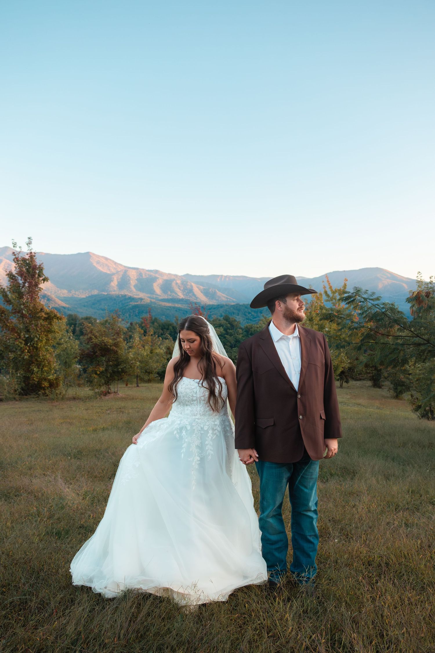 Newlywed couple in western attire standing together against a mountain landscape at sunset.