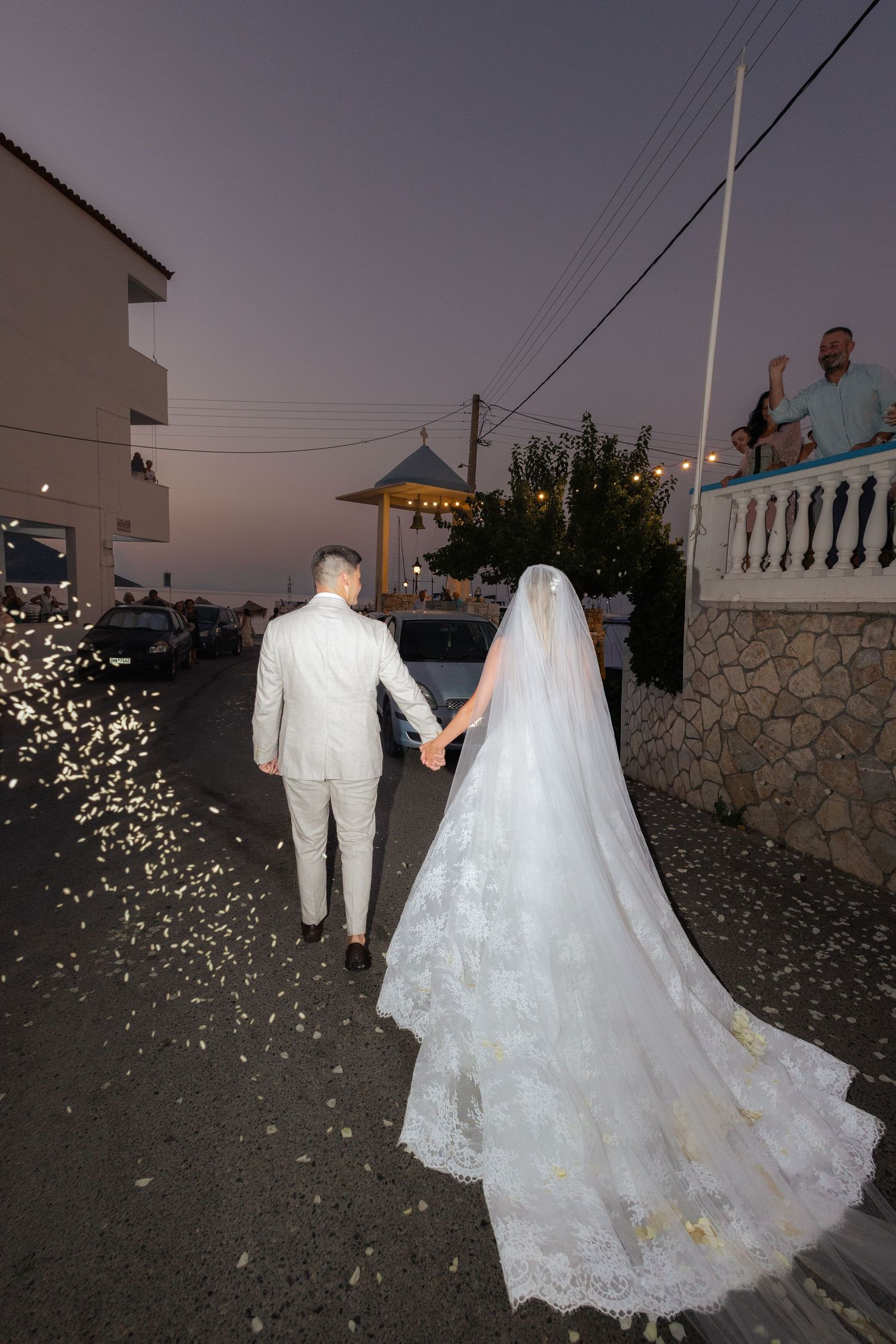 A newlywed couple walks along a stone path at dusk with confetti falling around their elegant white wedding attire.