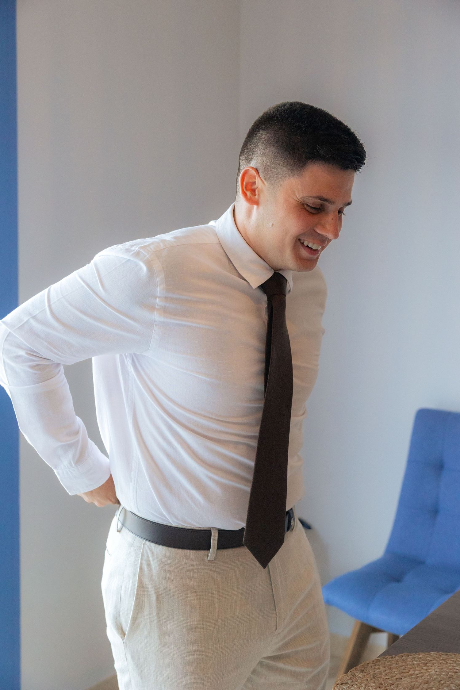 Professional wearing white dress shirt and black tie leans over while smiling in an office setting with blue chairs.