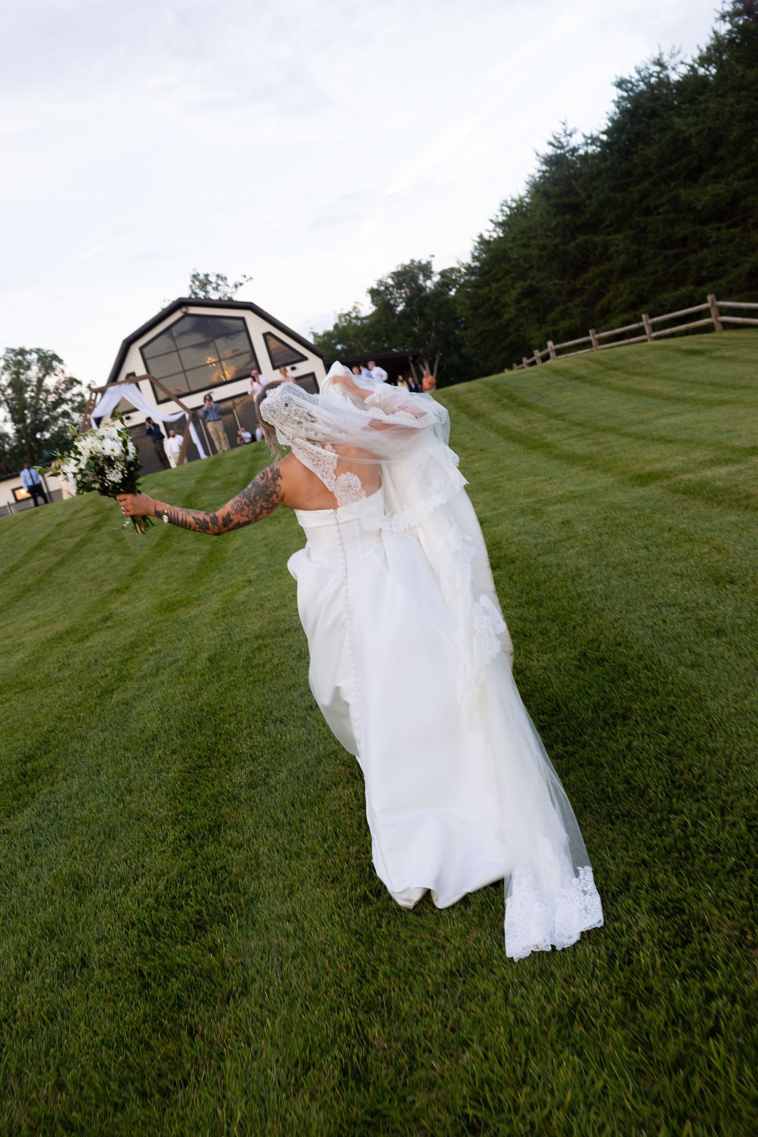 A bride in a flowing white dress leans back dramatically on a grassy hillside with a barn in the background.