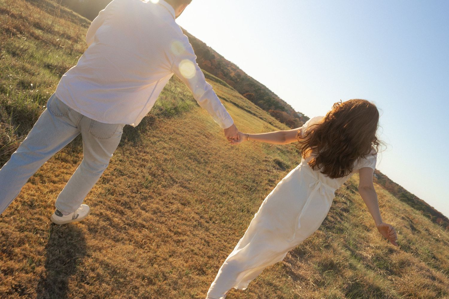 Two people in white outfits hold hands while spinning in a golden field at sunset.