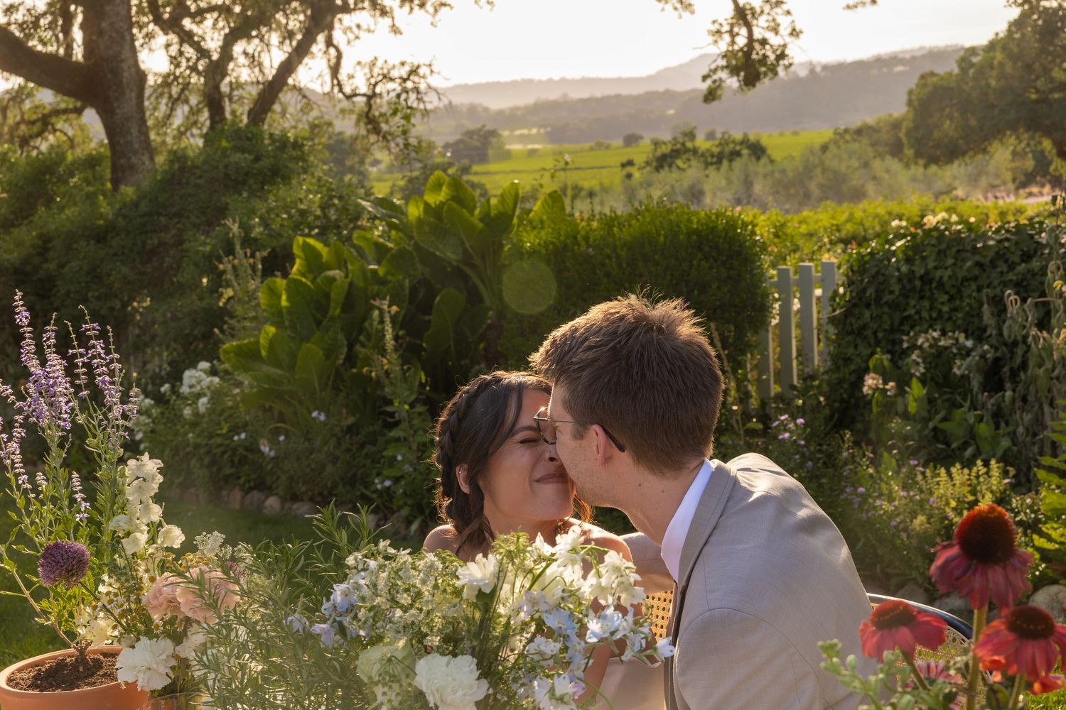 Couple shares romantic moments at sunset in a garden filled with blooming flowers and lush greenery.