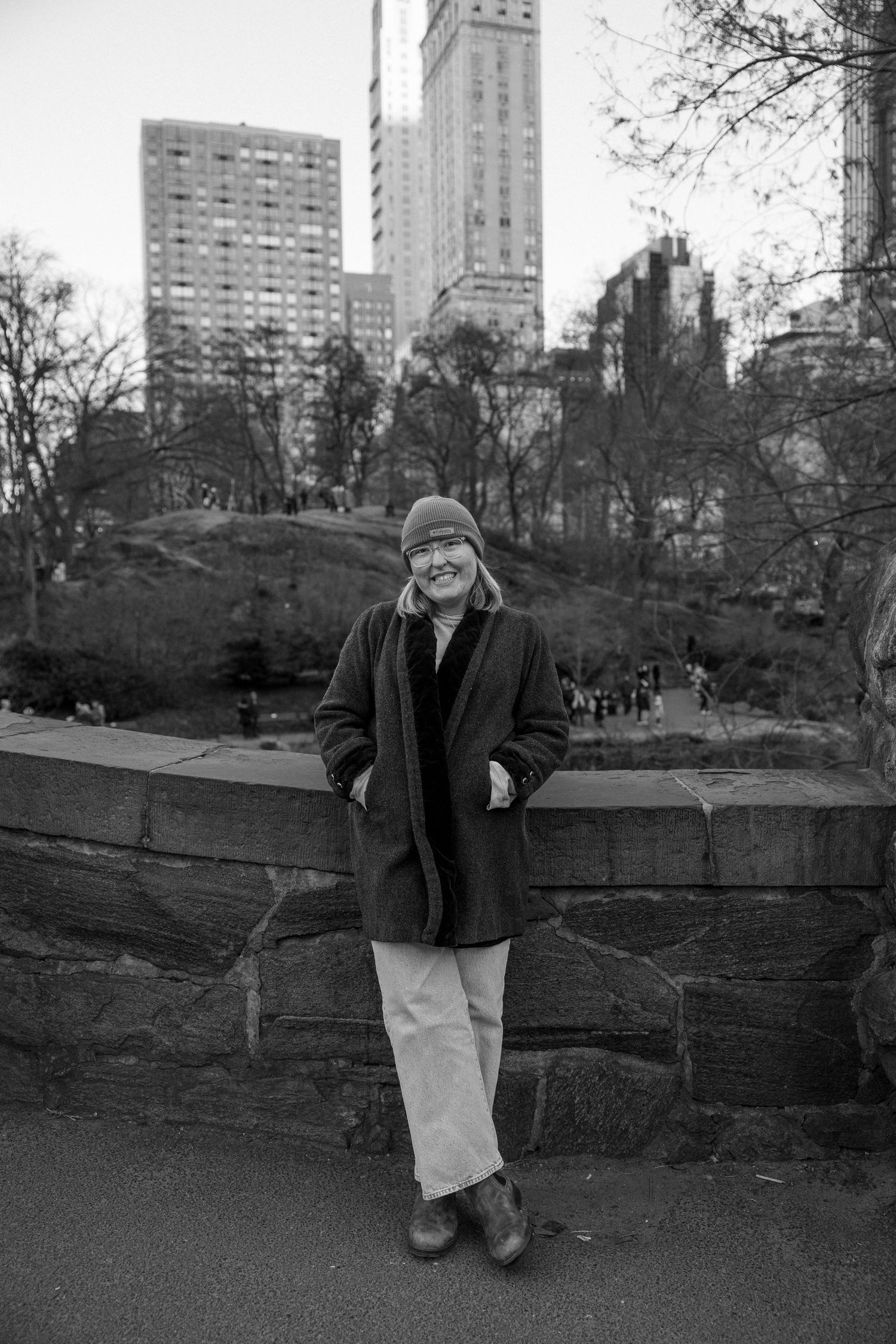 Black and white photo of someone in winter attire leaning against stone wall in Central Park with NYC buildings in background.