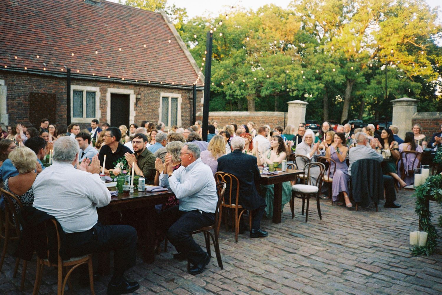 Wedding reception tables set up outdoors with string lights and brick architecture in the background.