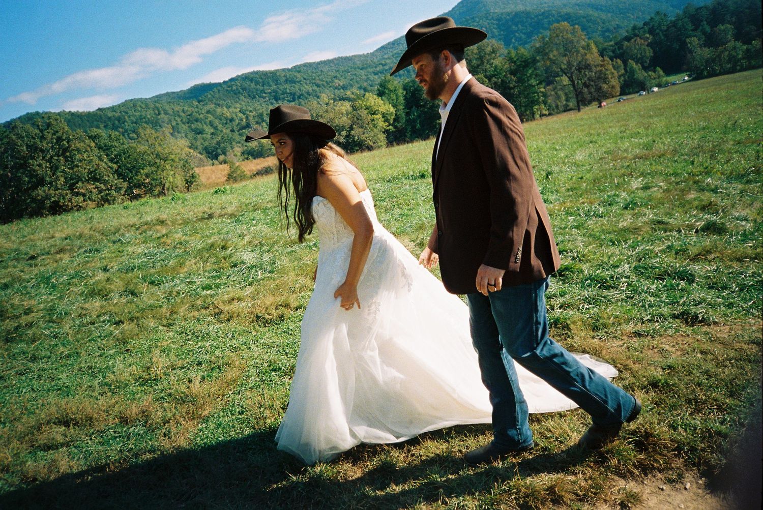 A couple walks through a scenic mountain meadow on a sunny day with green rolling hills in the background.