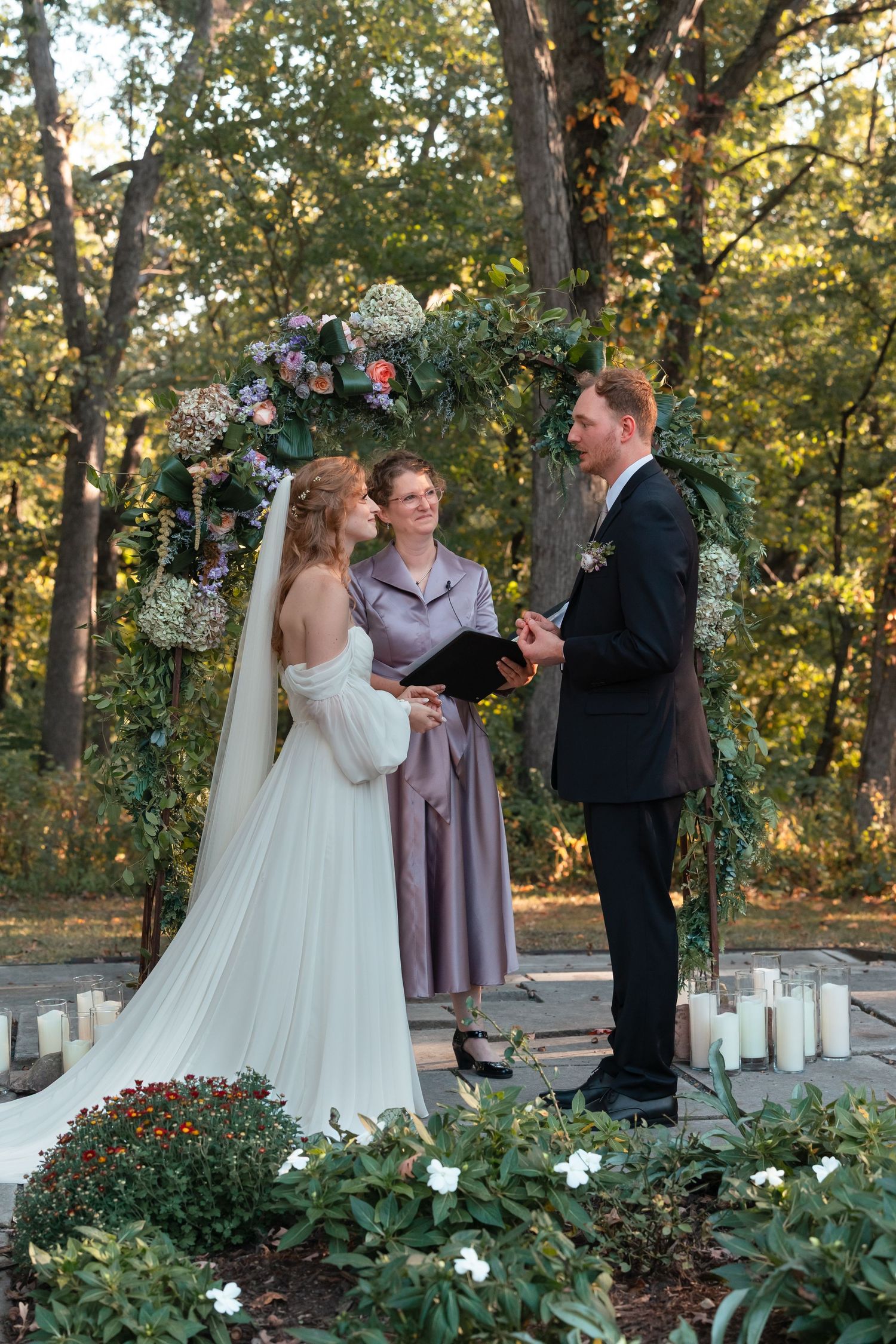 Wedding ceremony taking place outdoors with floral decorations and trees in golden sunlight.
