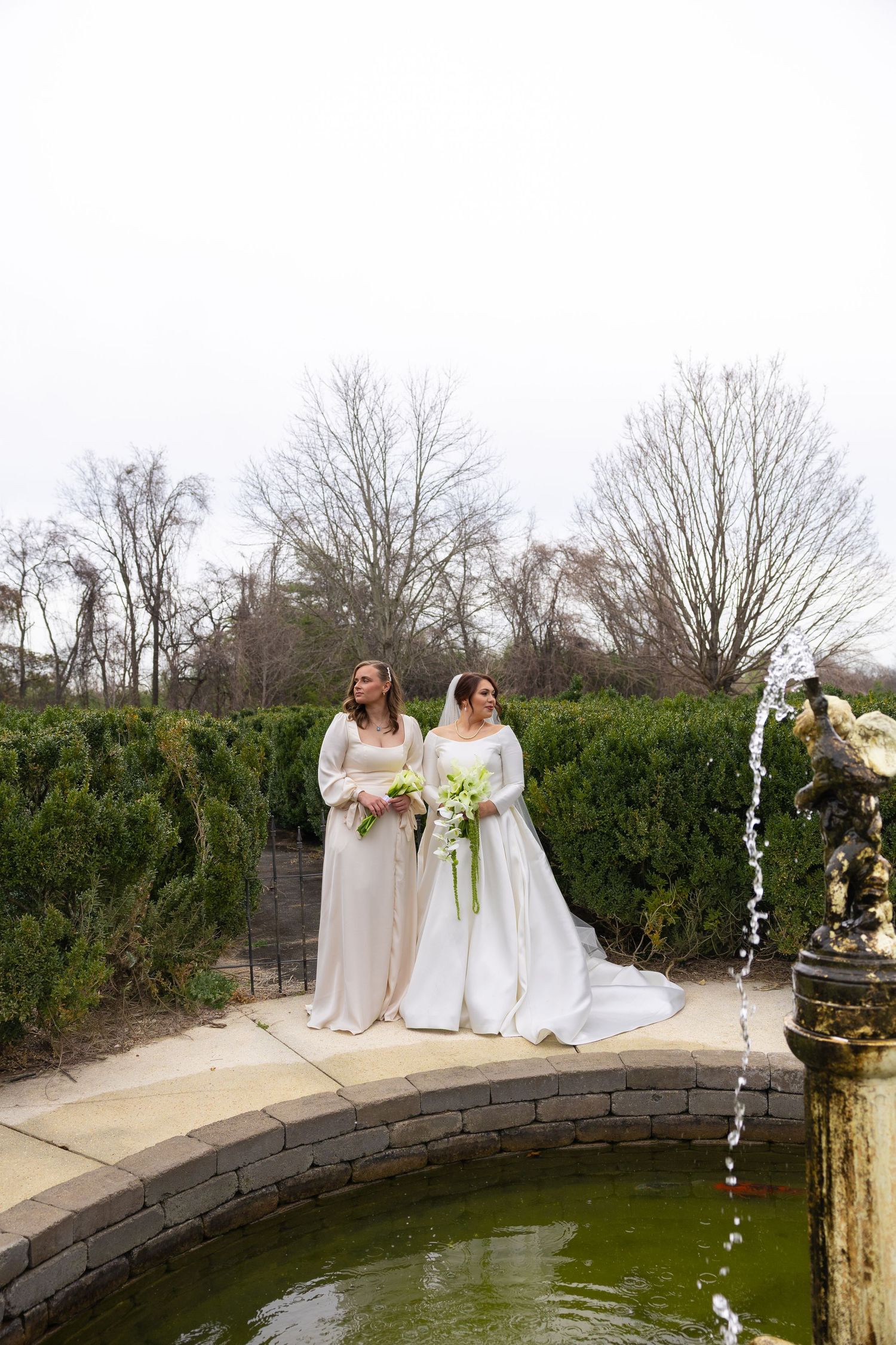Wedding party in white dresses posing by a curved stone wall with a fountain in a garden setting.