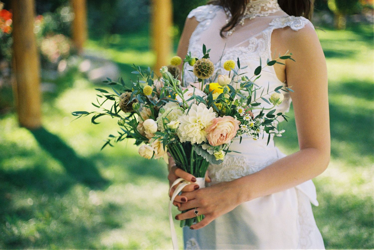 A delicate bridal bouquet with white and yellow flowers held against a white dress in sunlit garden.