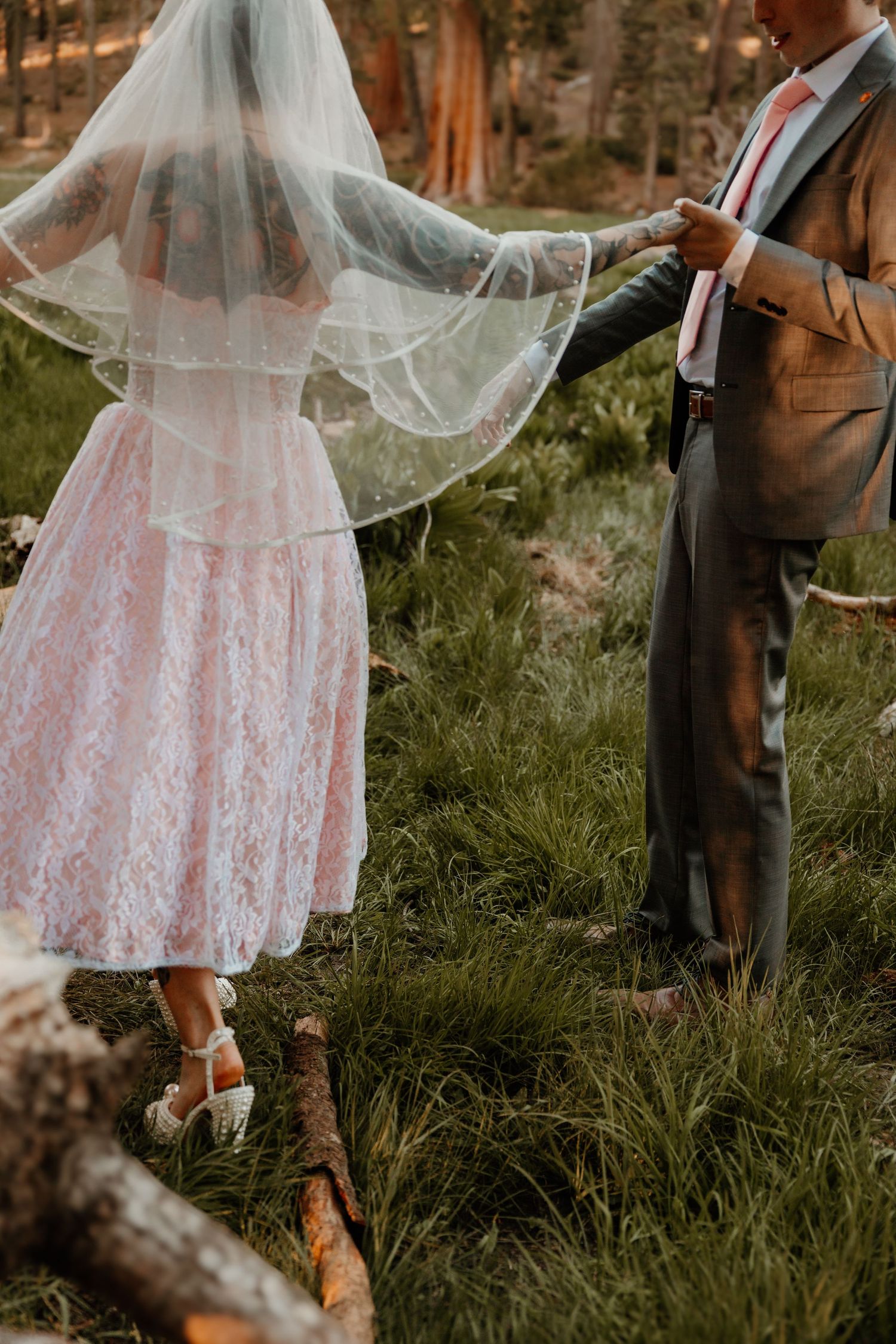 Bride in pink lace dress and flowing veil poses with groom in meadow surrounded by pine trees.