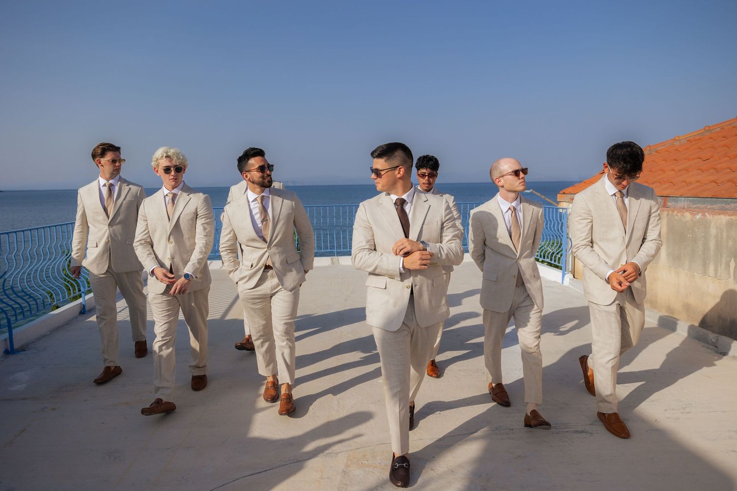 Groomsmen in light beige suits pose together on a Mediterranean seaside terrace.