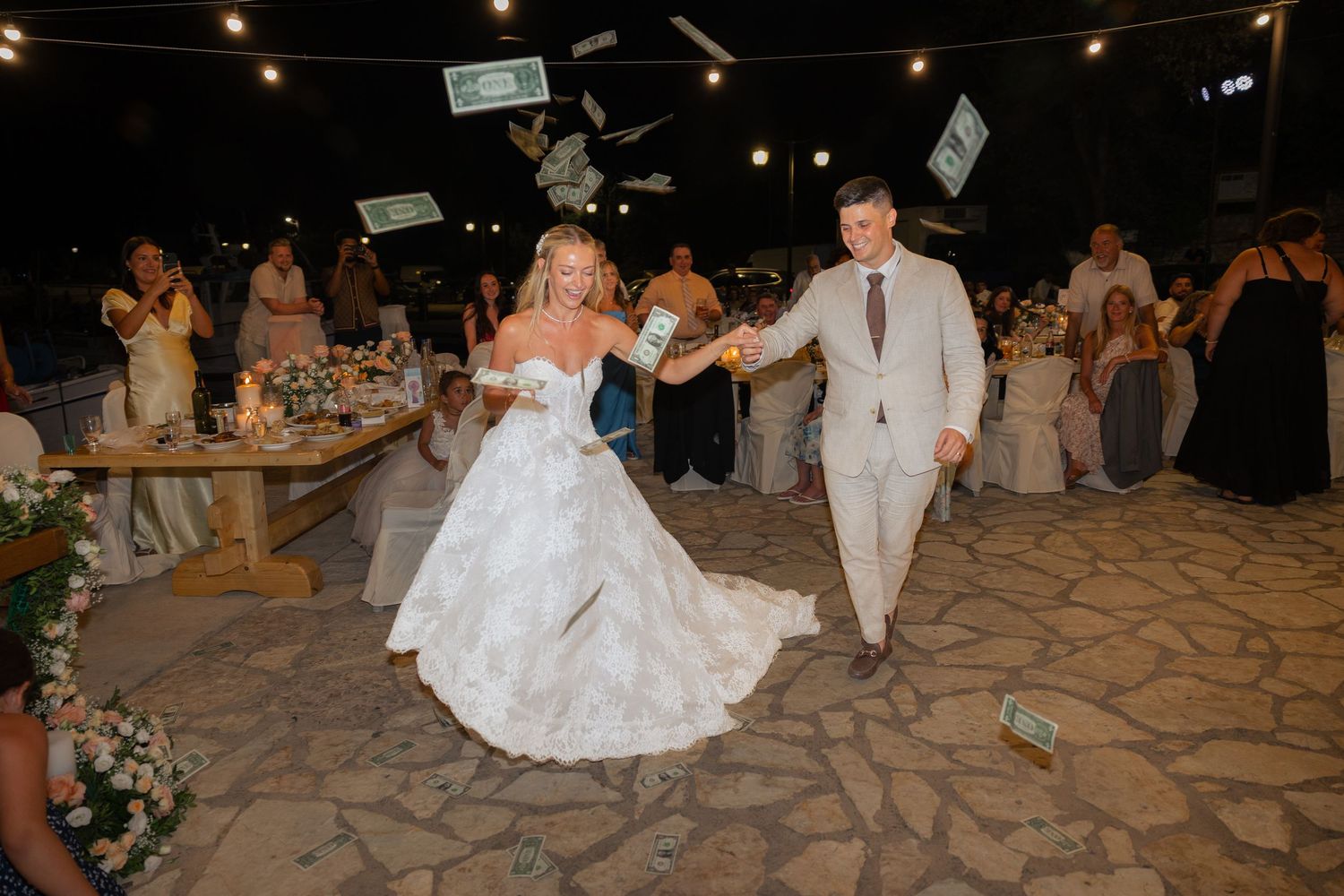 Wedding guests toss money in celebration as newlyweds dance on a stone patio during an evening reception.