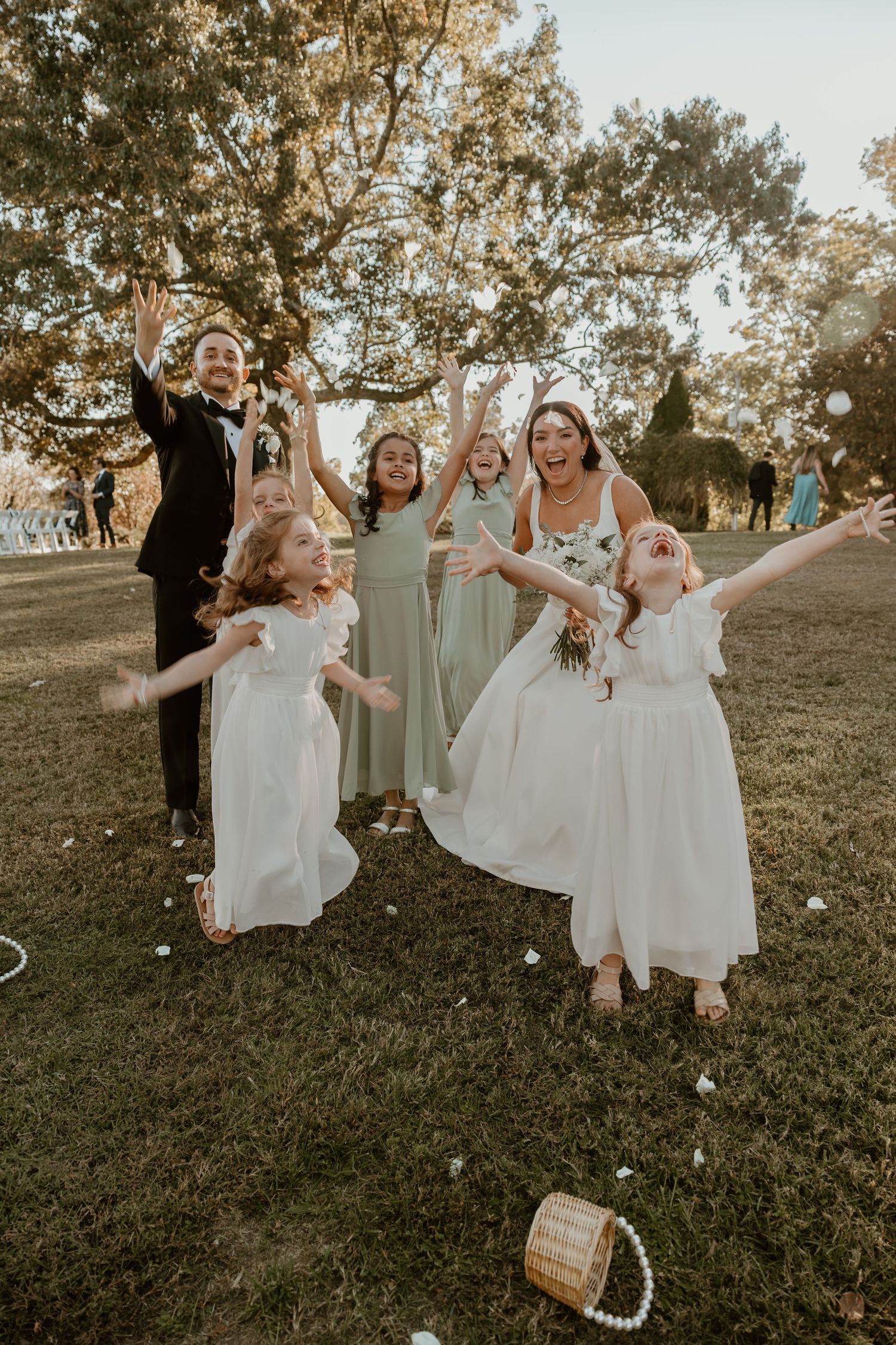 A joyful wedding party celebrating on a lawn with flower girls in white dresses.