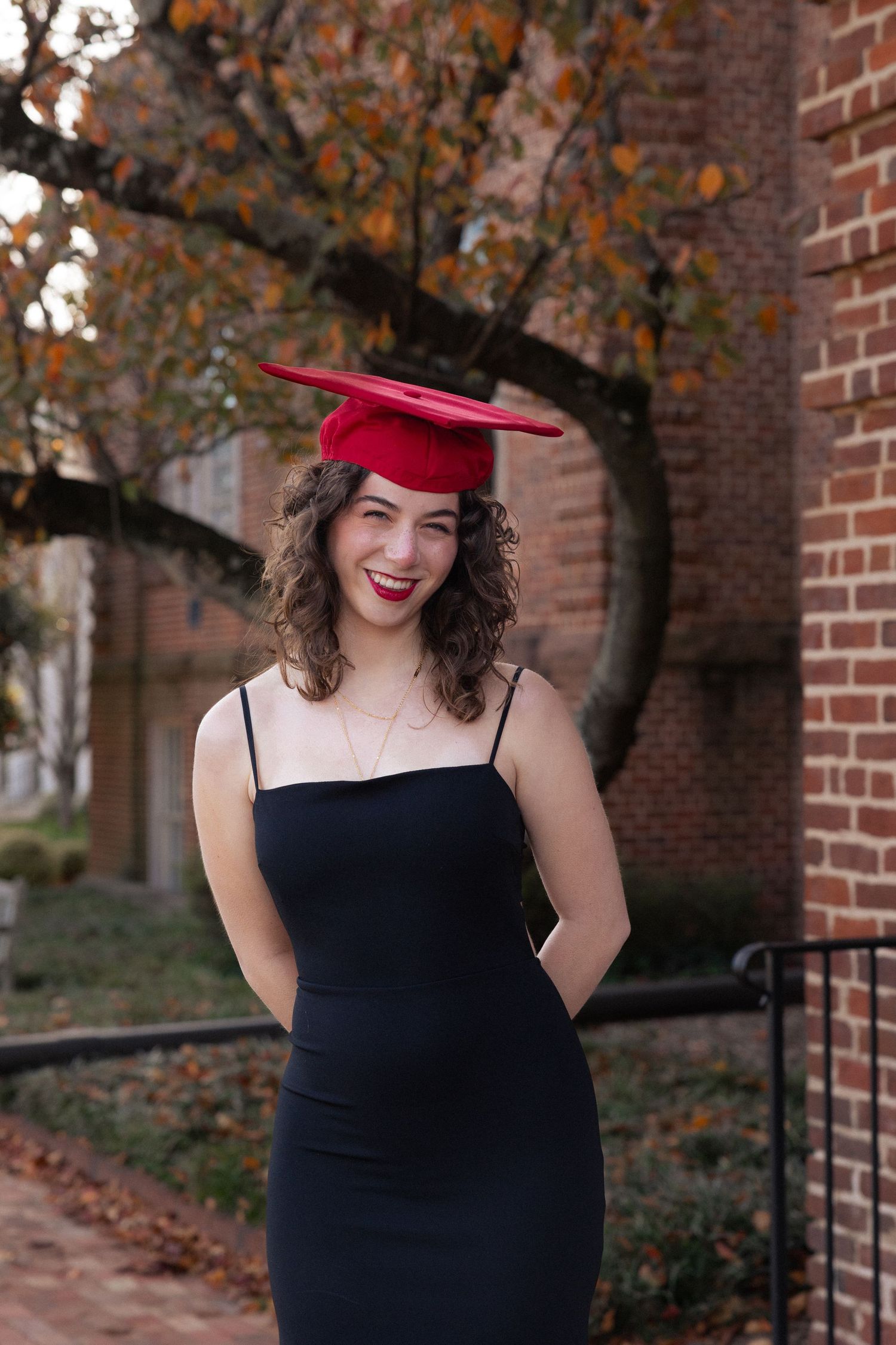 Smiling graduate in black dress and red mortarboard cap poses outdoors near brick building in autumn.