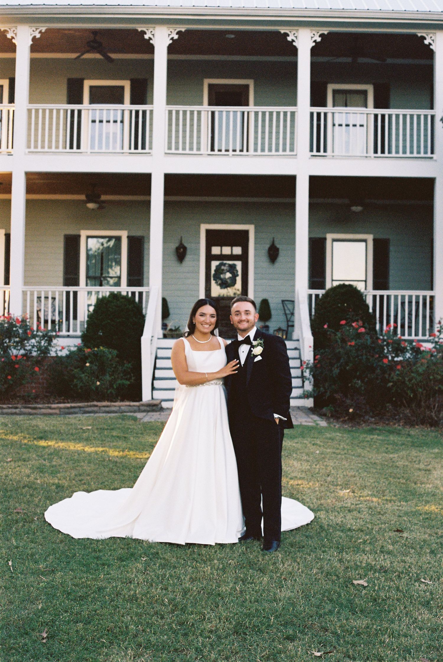 A couple poses in wedding attire in front of a charming two-story house with white columns and balconies.