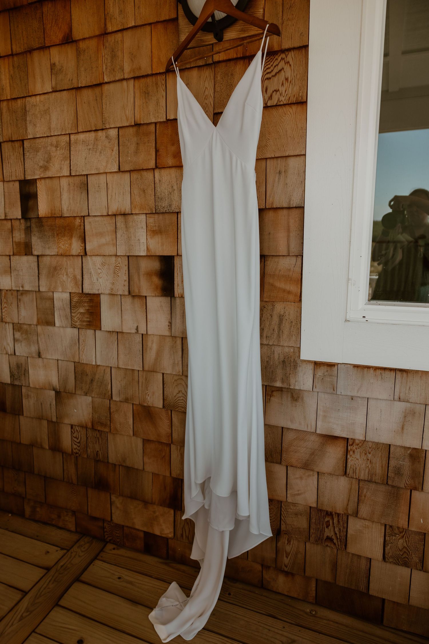 Elegant white wedding dress hanging on cedar shingle wall near window.