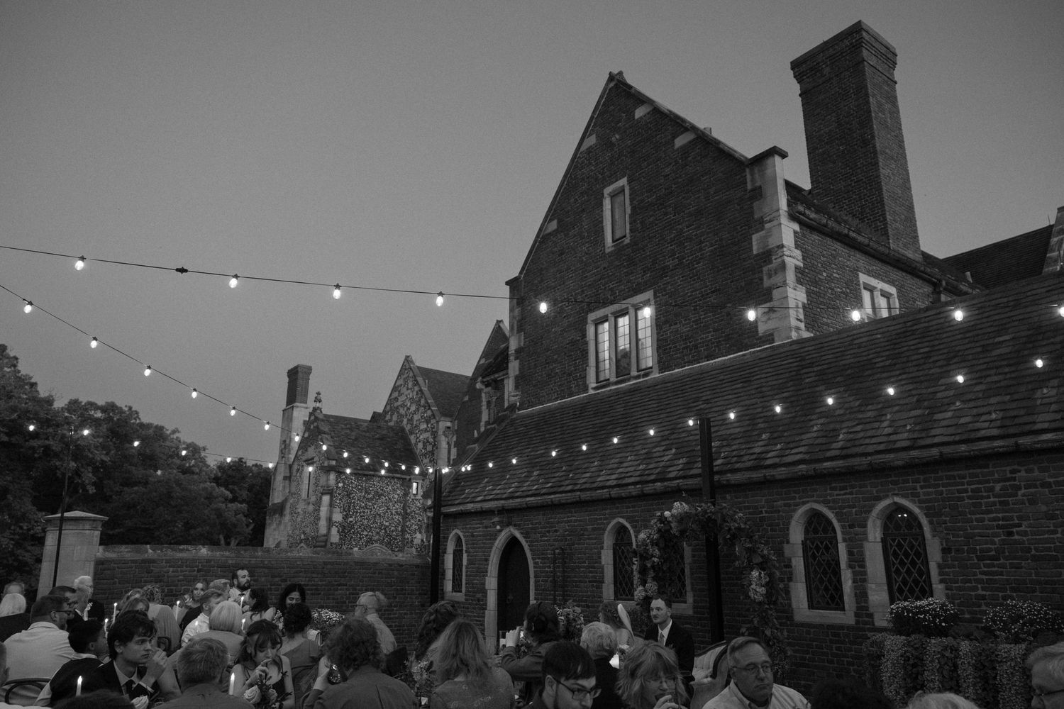 String lights illuminate an outdoor courtyard between historic brick buildings at dusk creating a cozy ambiance.