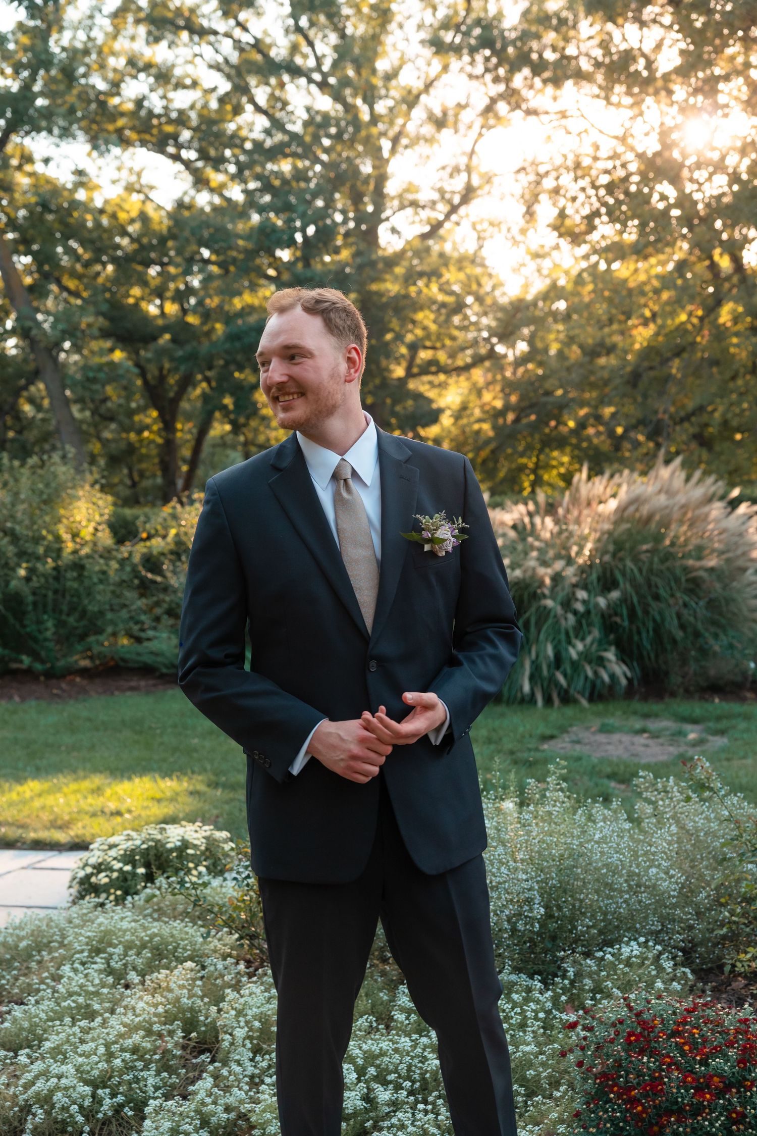 A groom in a navy suit and light tie smiles in a garden setting during golden hour.