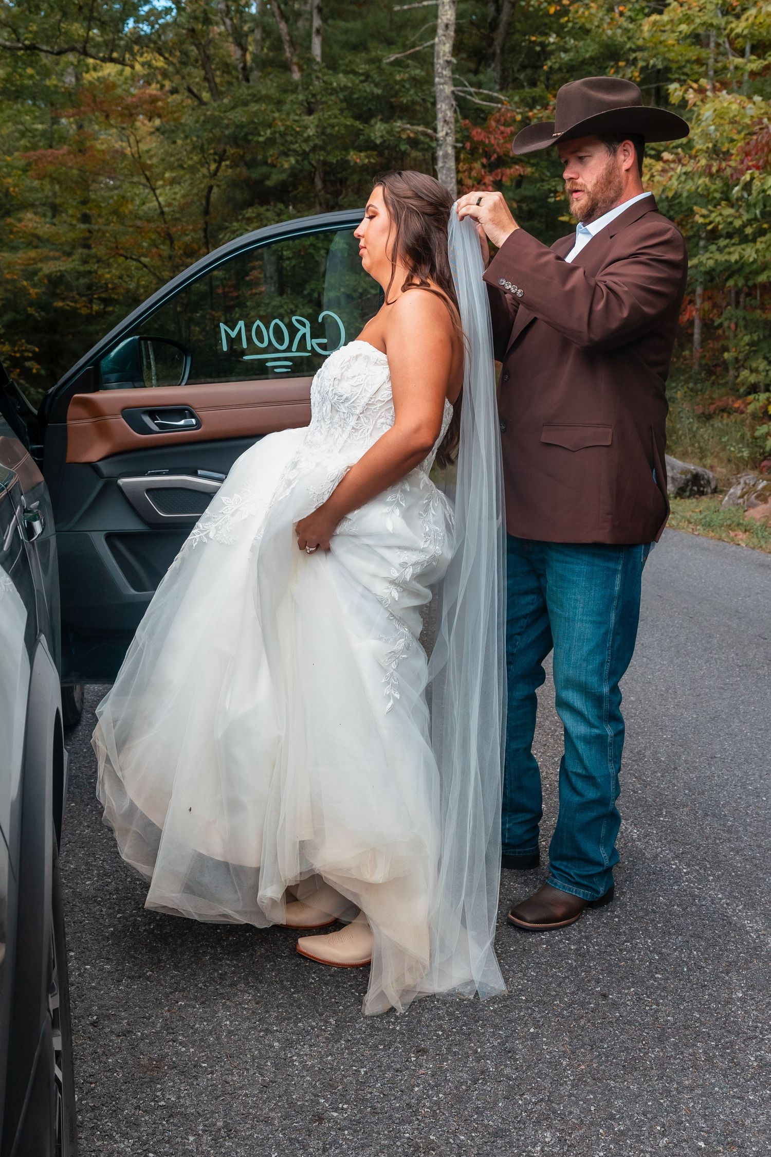 A couple in wedding attire standing by a car door in a wooded area during autumn.