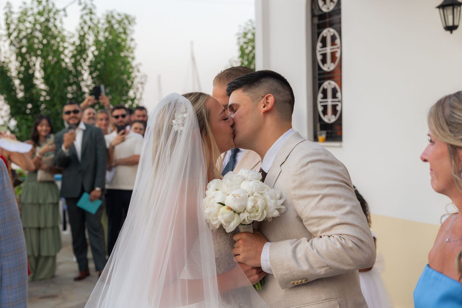 A wedding couple shares a romantic kiss while holding a white bouquet during an outdoor ceremony.
