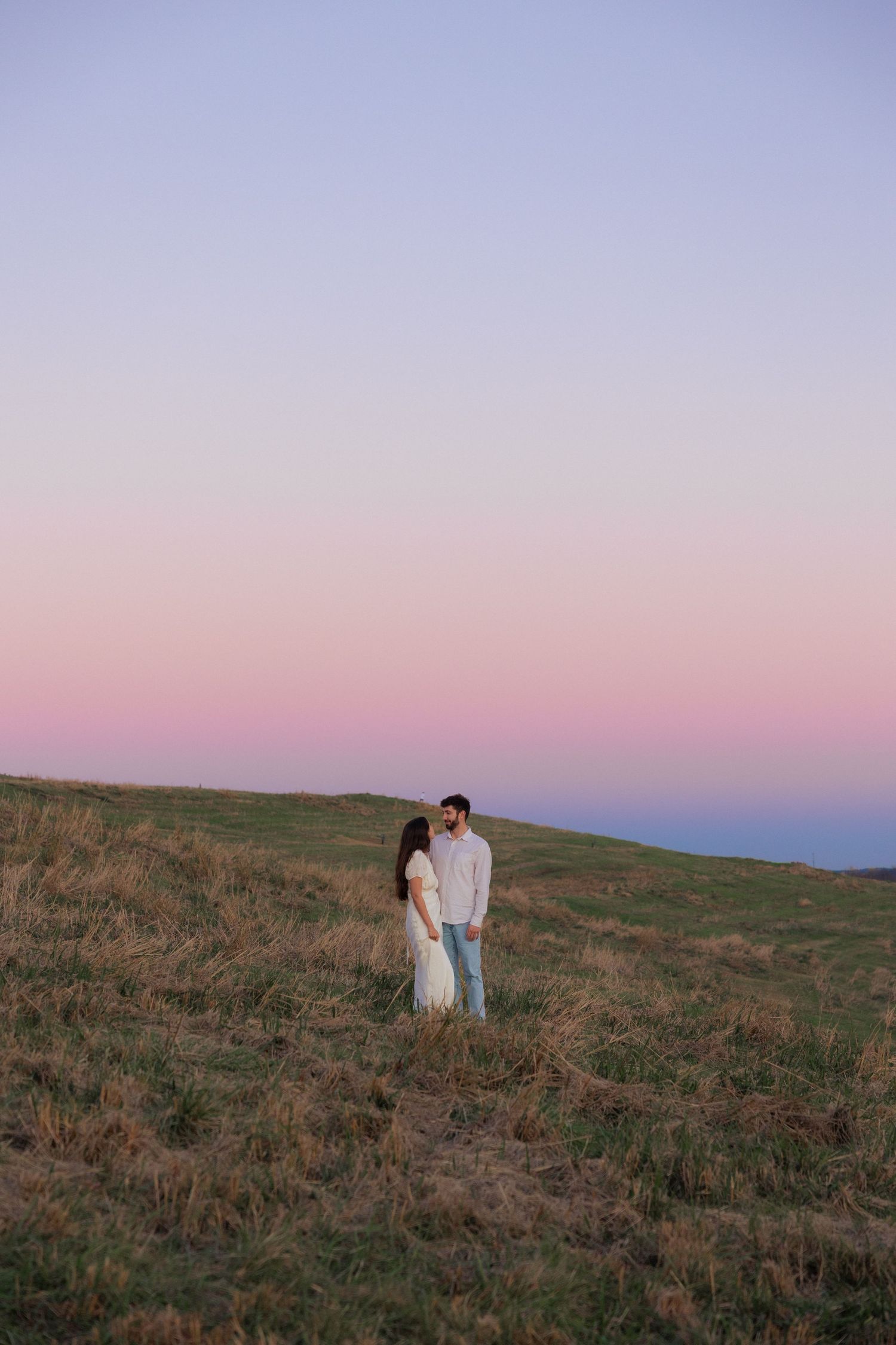 Two figures in white clothing stand together on a hilltop against a dusky purple sky.