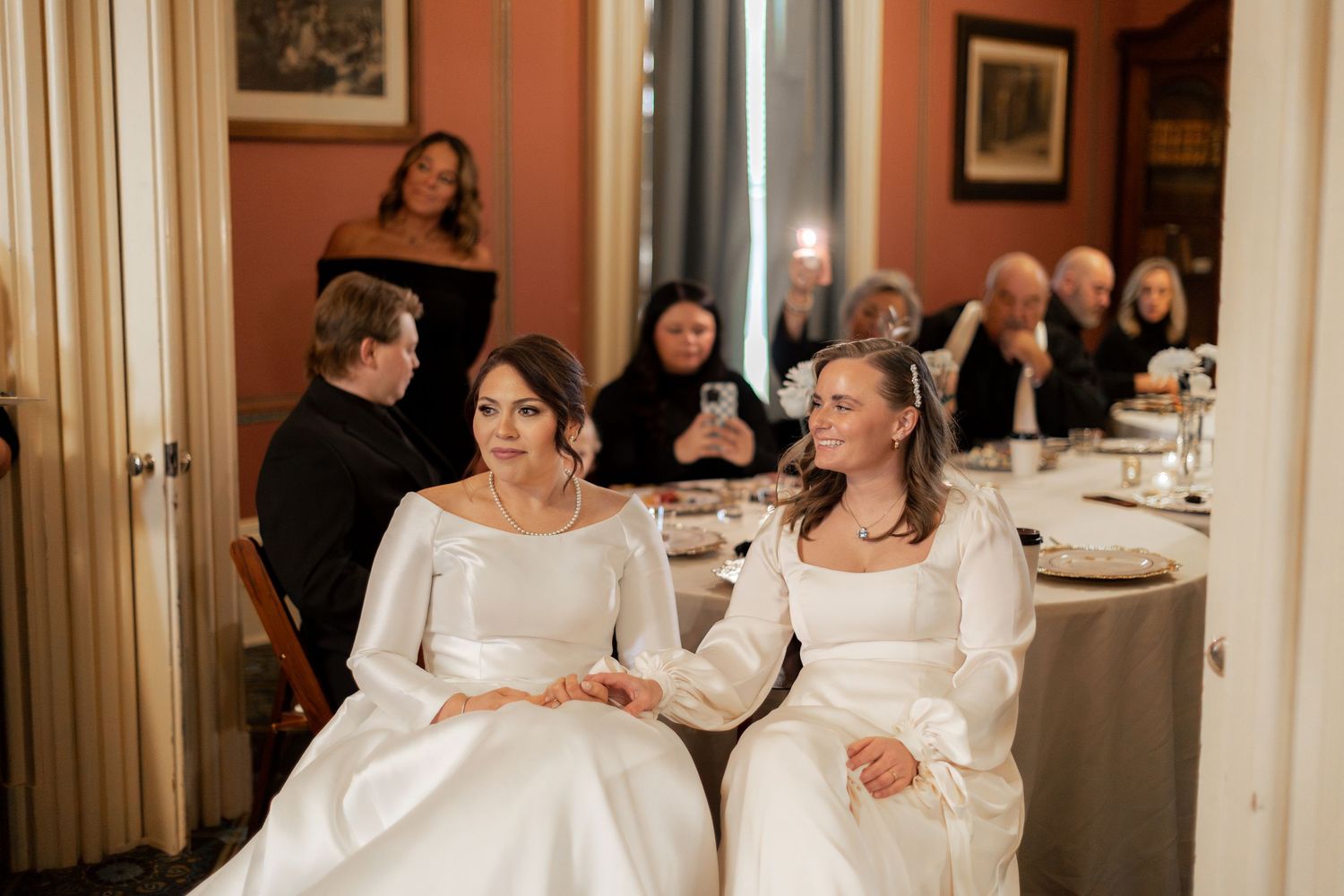 Two people in white dresses sit together at an indoor wedding reception dinner table.