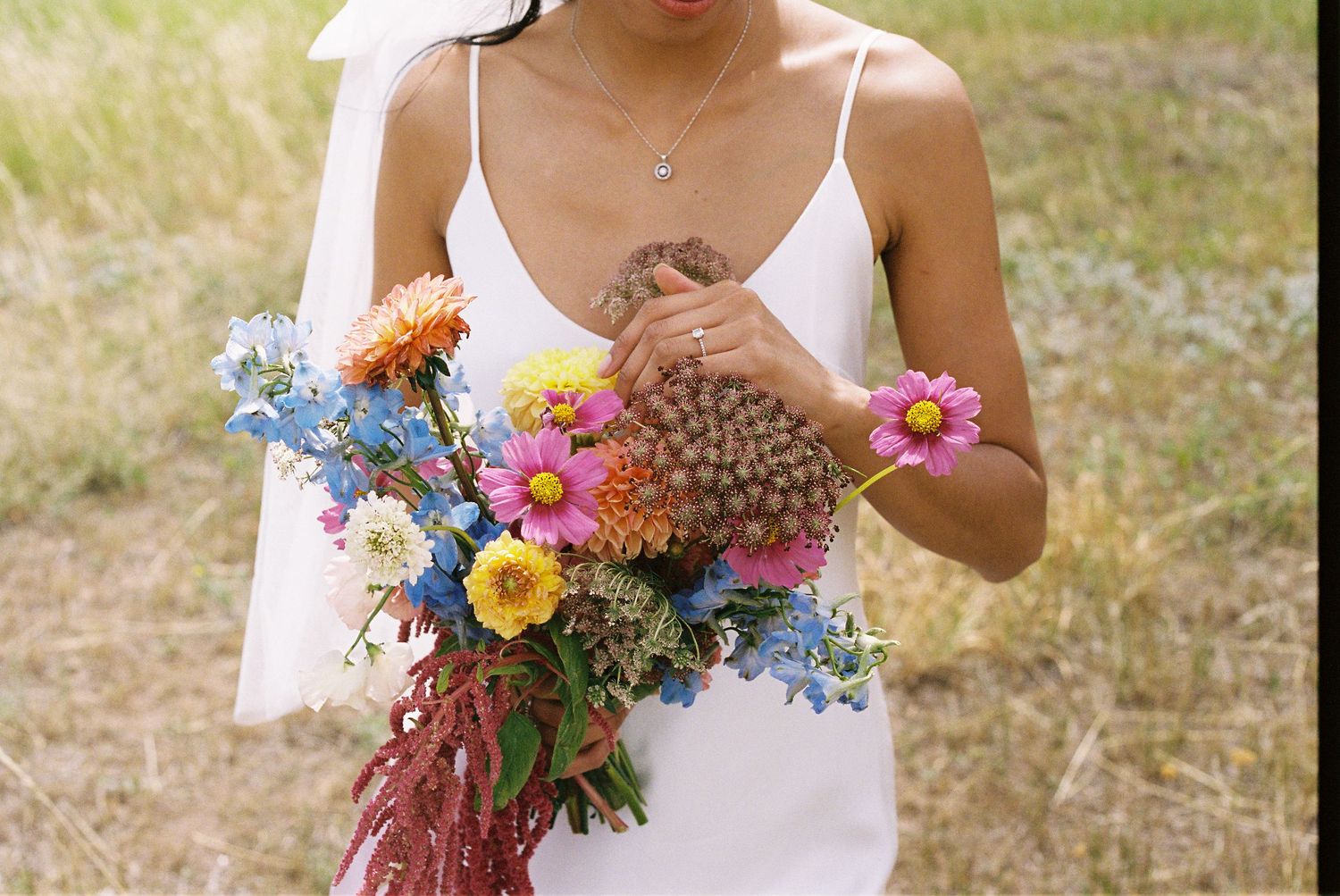 A colorful summer bouquet with orange, yellow and blue wildflowers held against a white dress in a field.