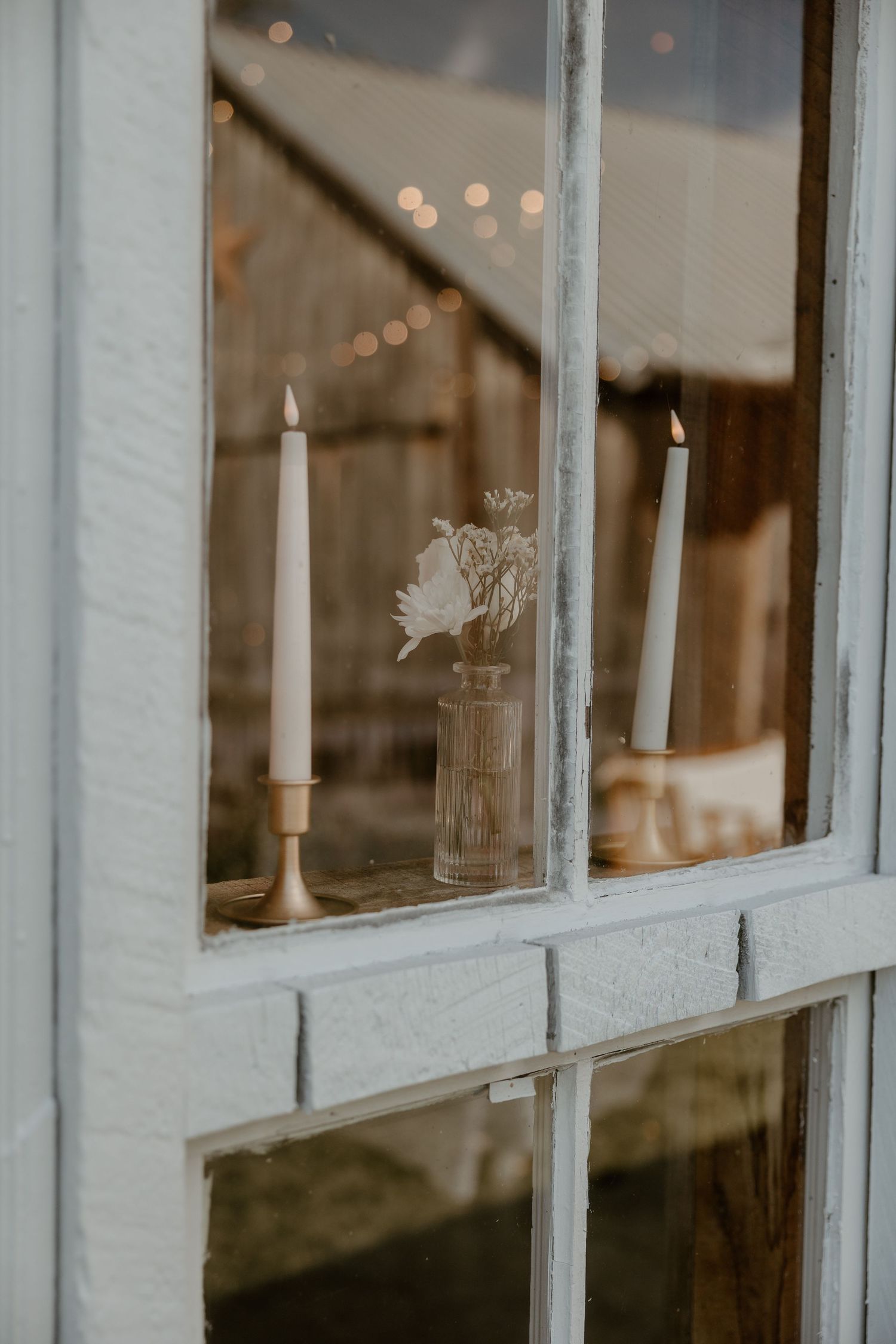 White taper candles and delicate flowers in a glass vase sit on a windowsill with soft bokeh lights in background.