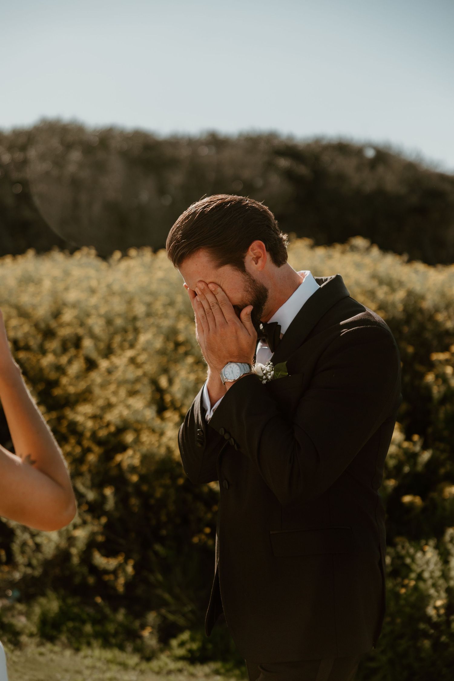 A groom becomes emotional and covers his face during an outdoor wedding ceremony.