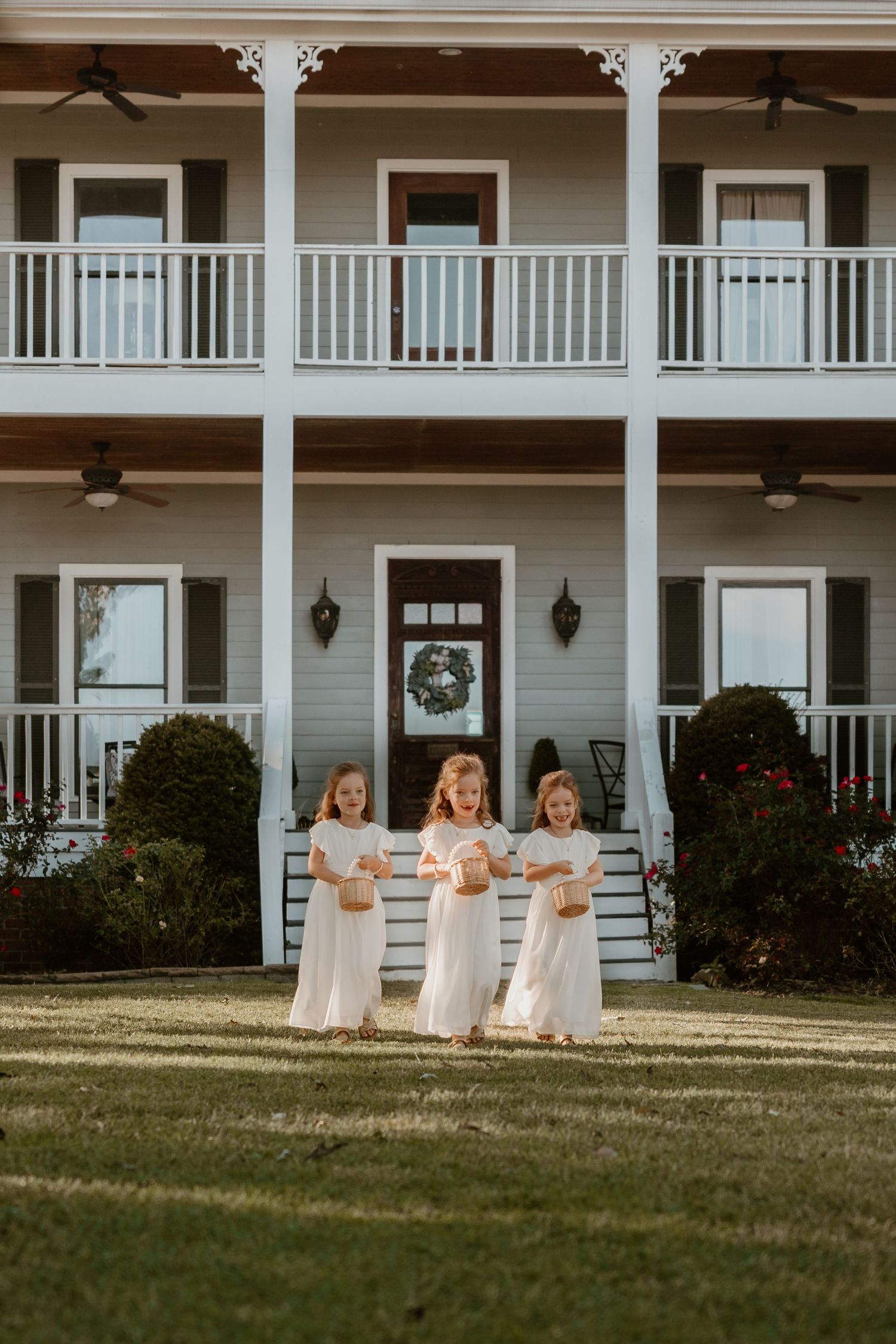 Three young flower girls in white dresses stand in front of a beautiful two-story colonial style house.