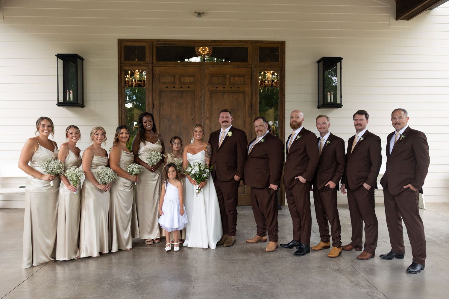 Wedding party lined up in formal attire with bridesmaids in light dresses and groomsmen in dark suits against white wall.